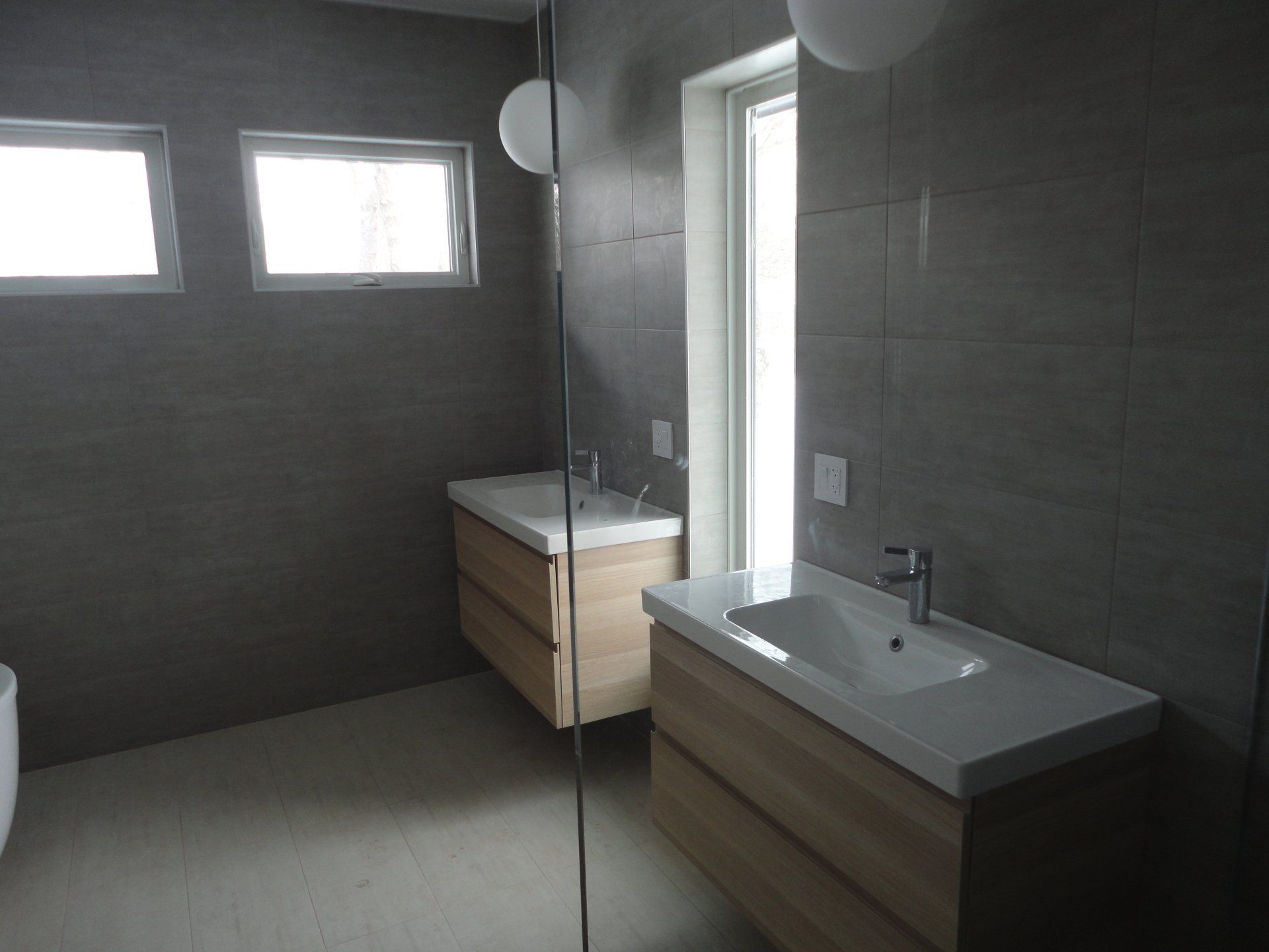 Modern bathroom with light wood vanities, white countertops, and grey tiled walls. Windows provide natural light.
