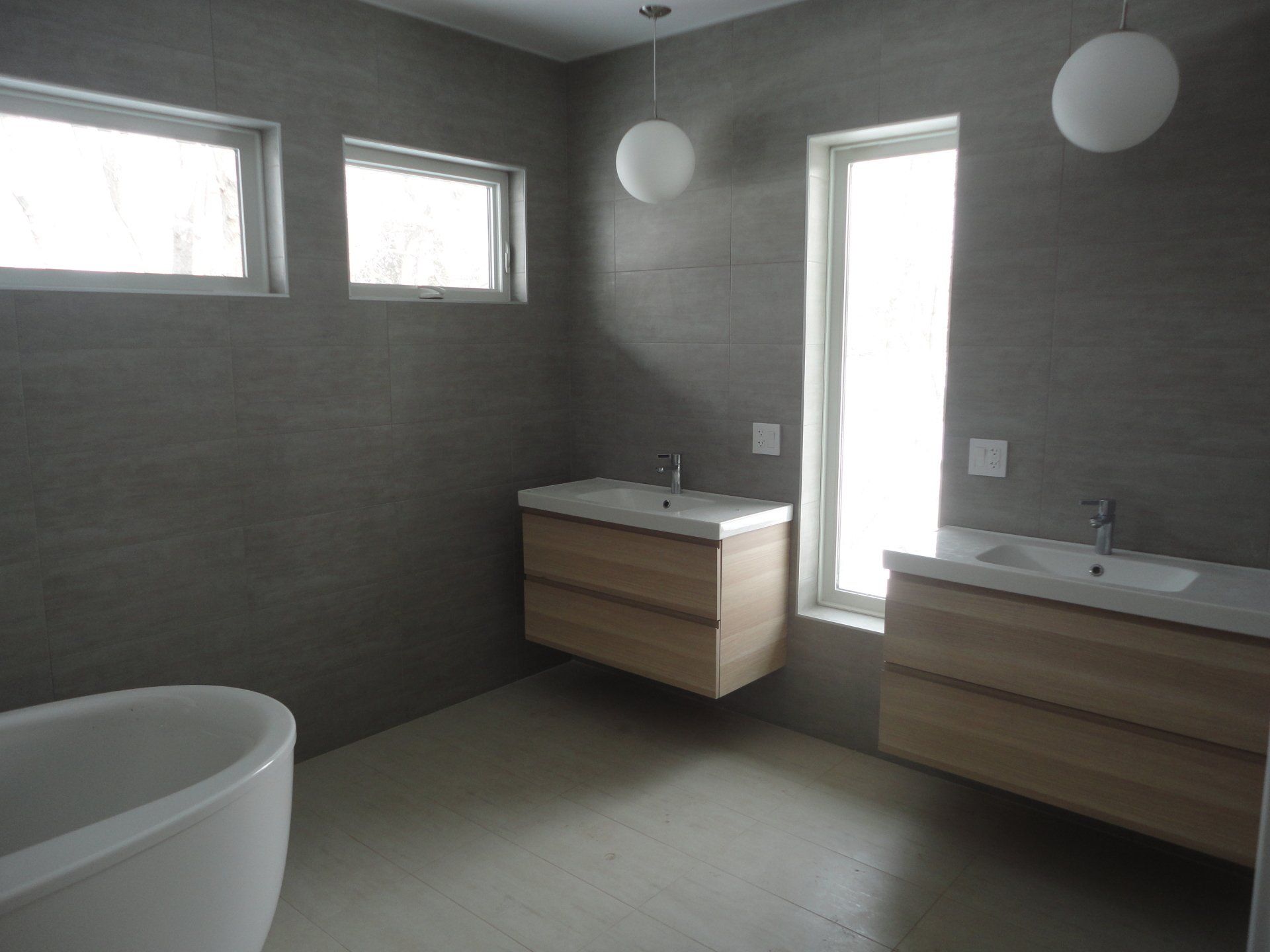 Modern bathroom with two wood vanities, large window, and soaking tub; gray walls and floor.