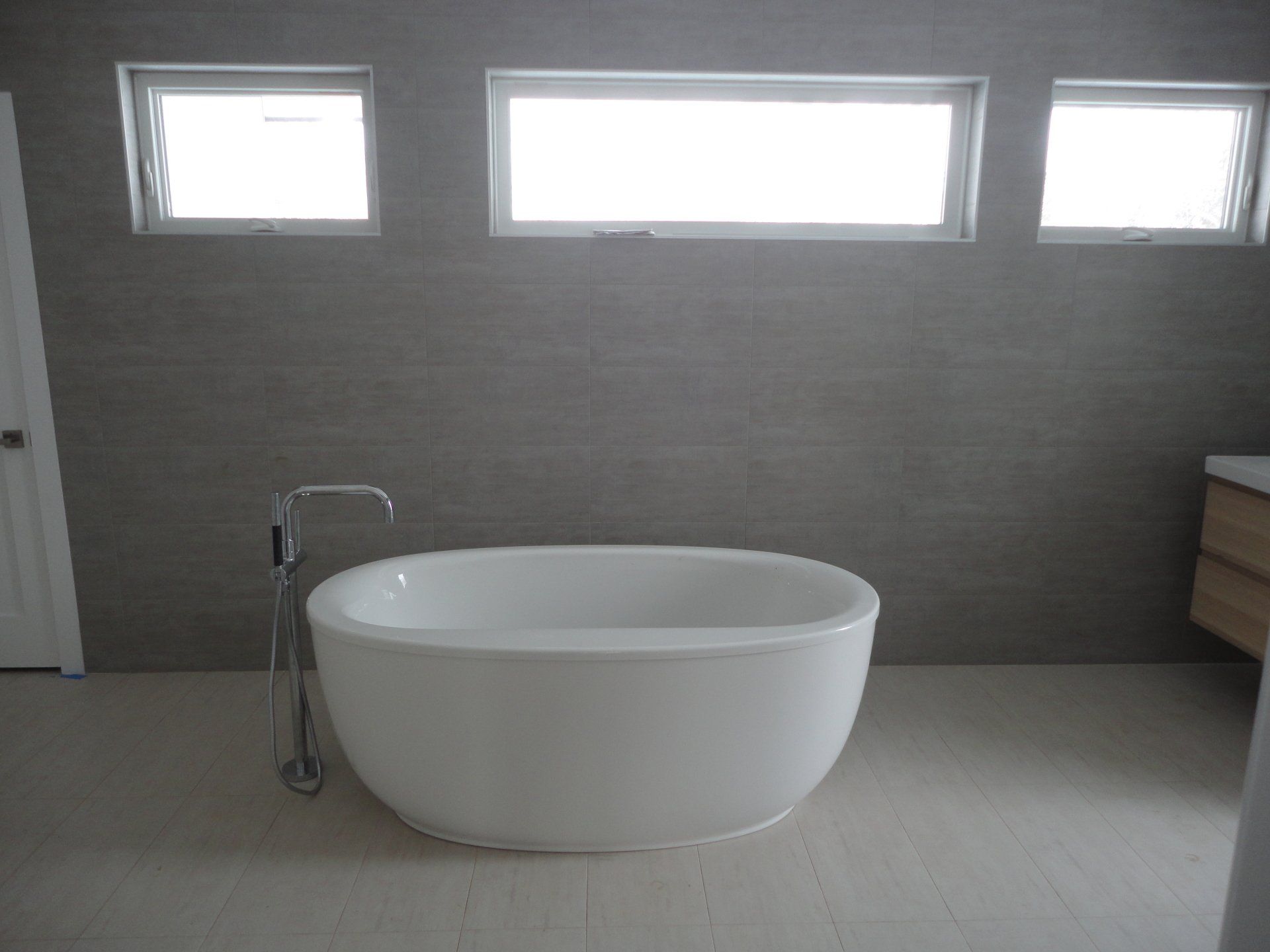 Freestanding white bathtub in a modern bathroom with gray tiled walls and three windows.
