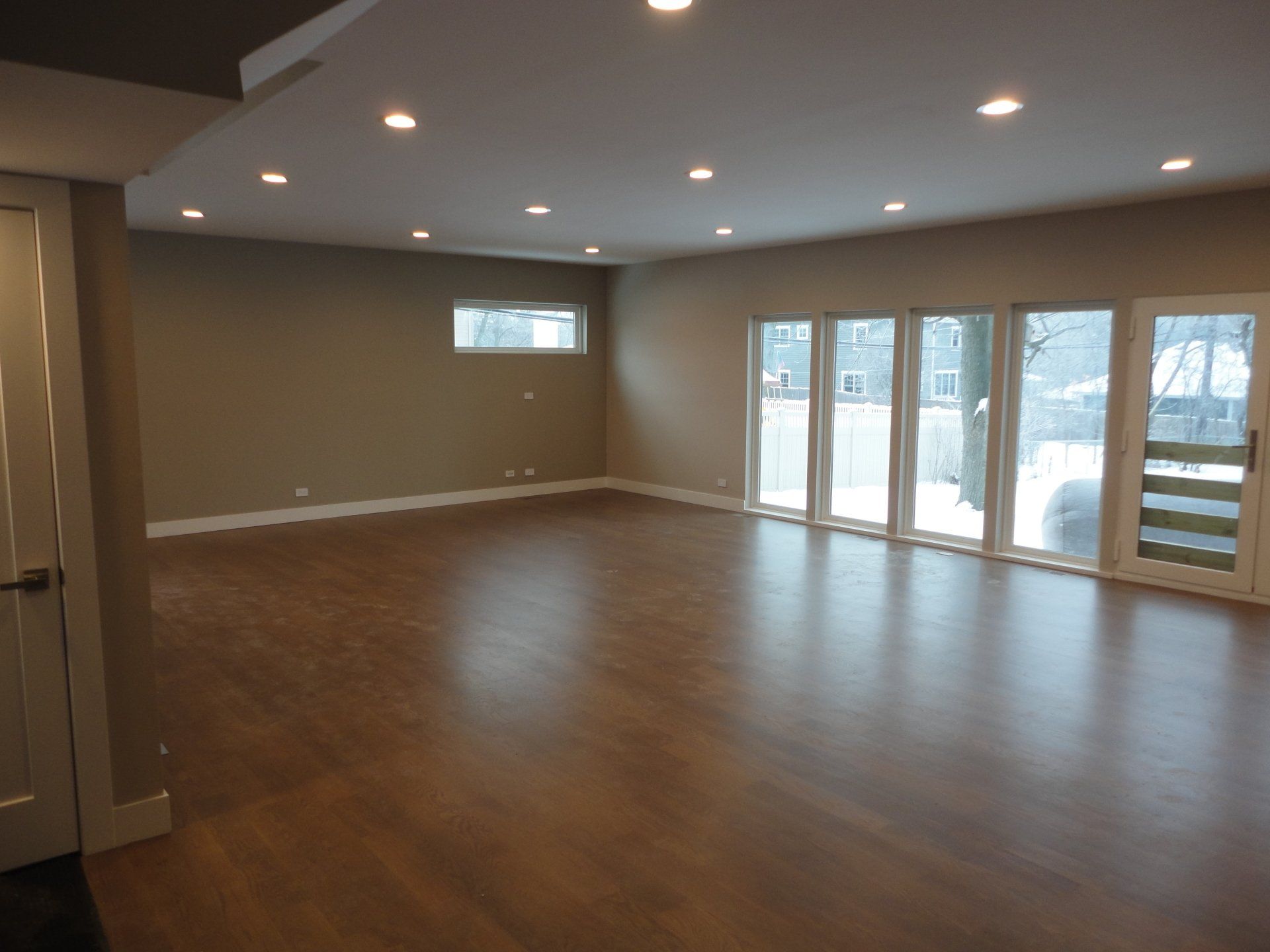 Empty living room with hardwood floor, large windows, and recessed lighting.