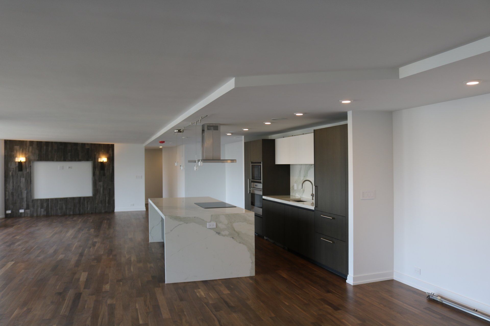 Modern kitchen with island, dark cabinets, and open living space; wood floors and accent wall.