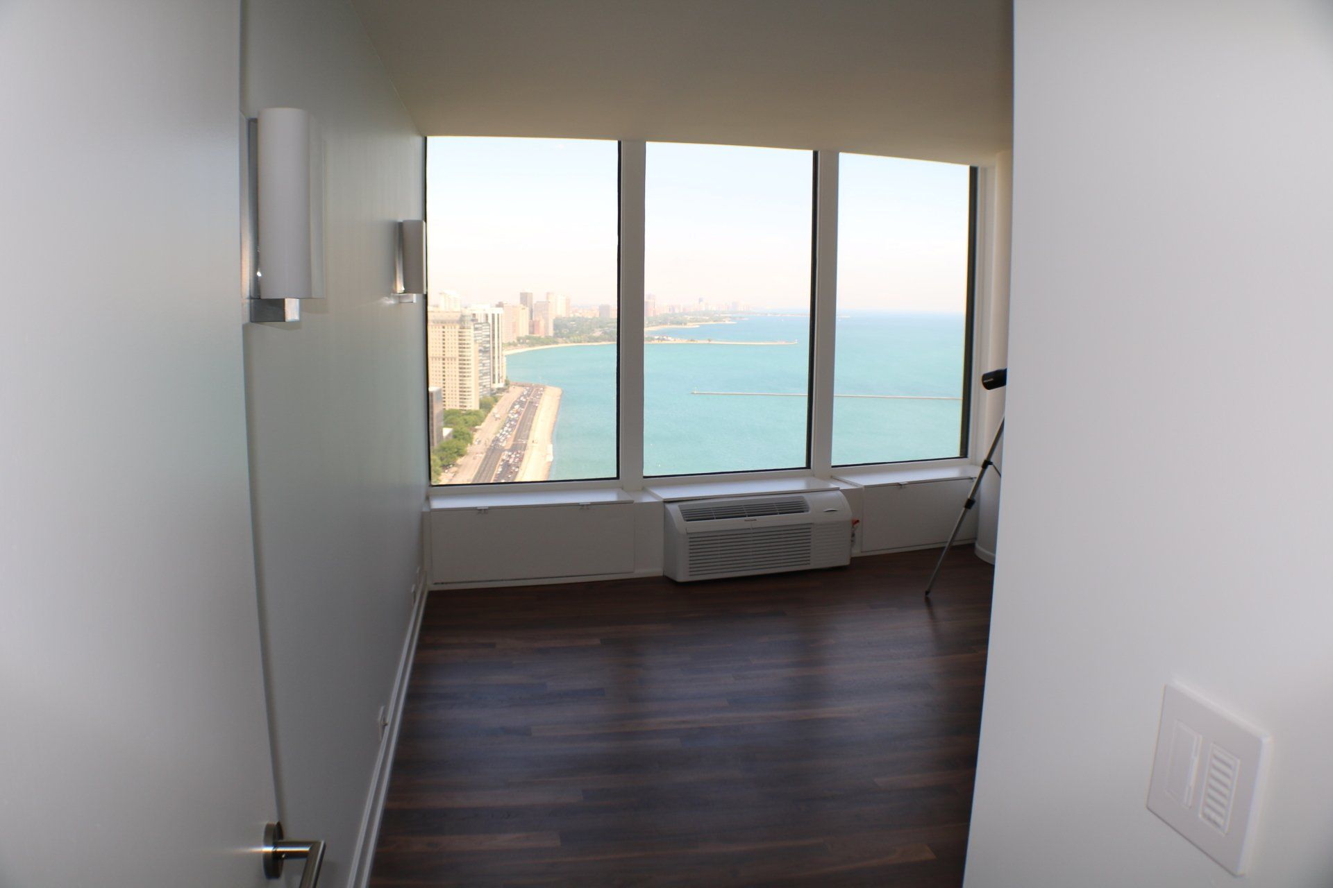Room with large windows overlooking city and water, dark wooden floor, white walls.