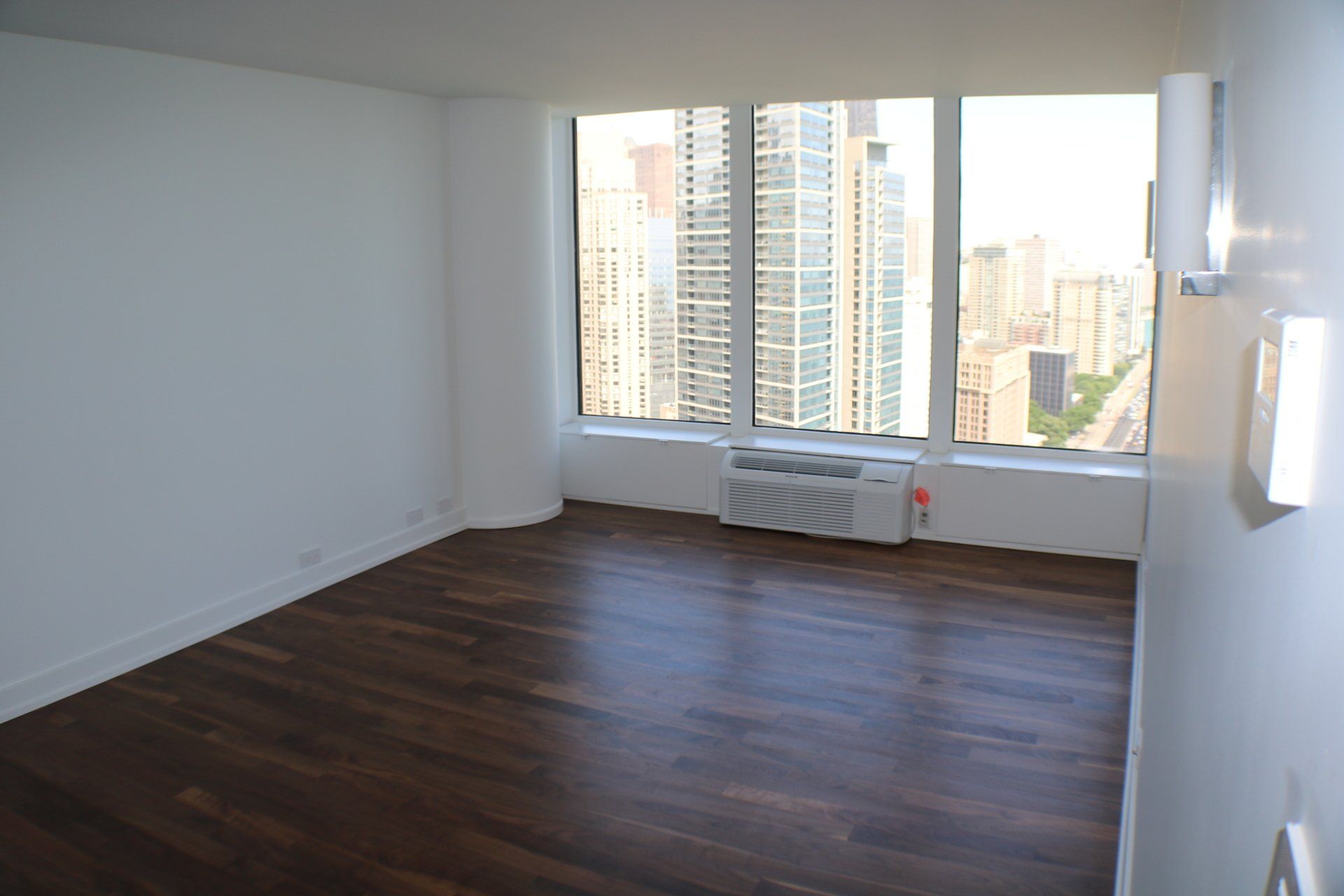 Empty room with dark wood floor, large windows overlooking city buildings, and two wall-mounted lights.