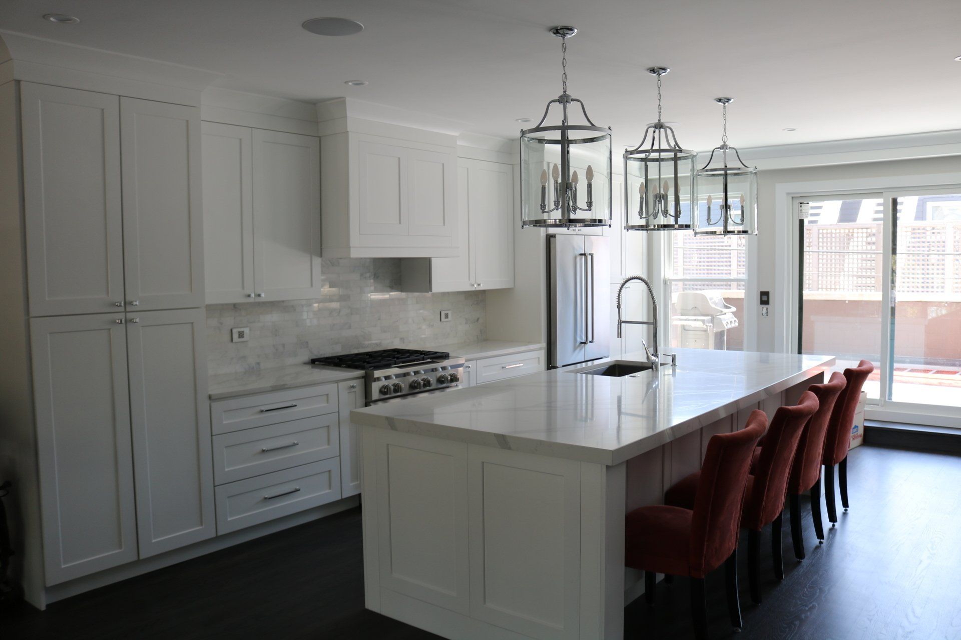 White kitchen with island, marble countertops, dark floors, red bar stools, and pendant lights.