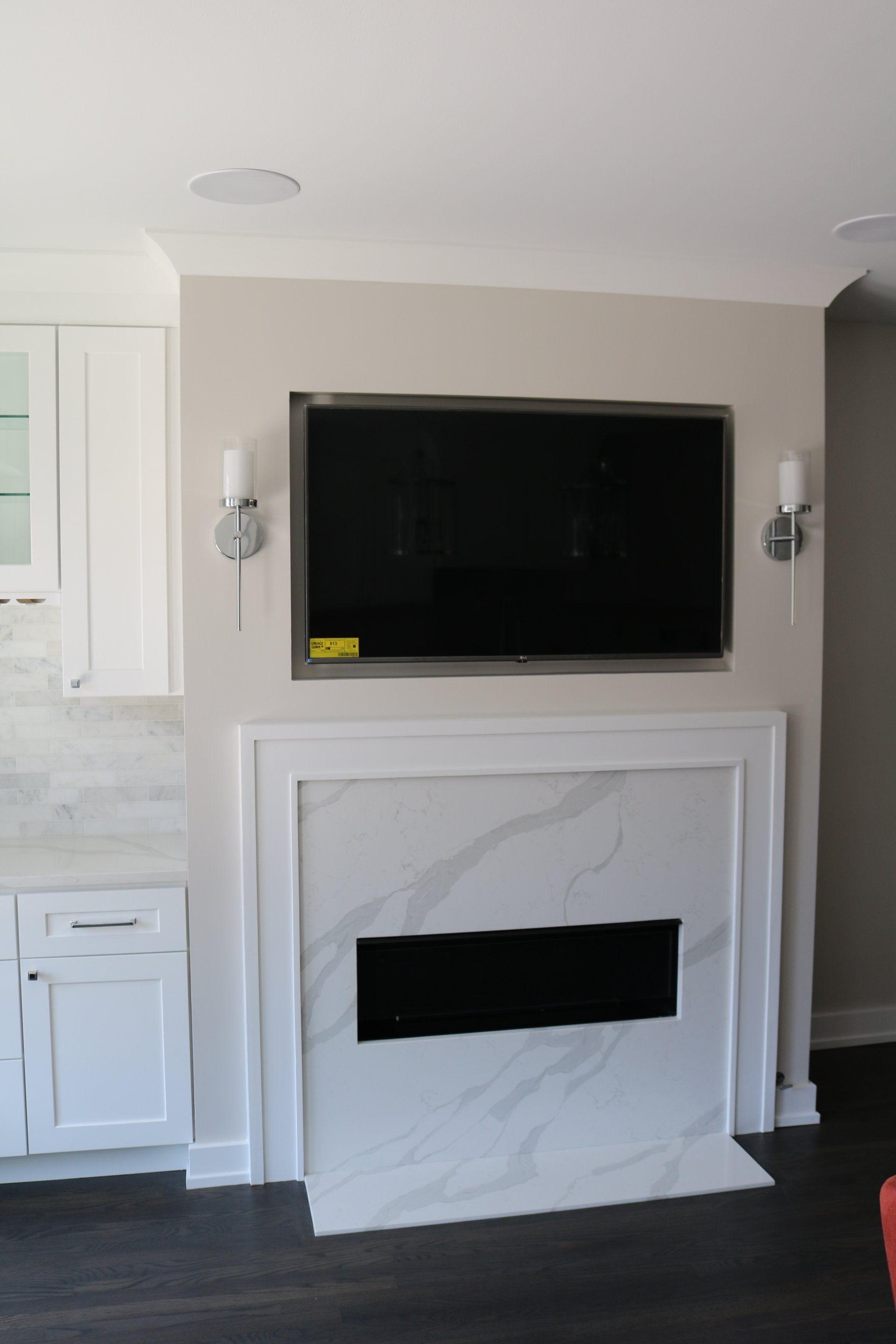 TV mounted above white marble fireplace with sconces. White cabinets and dark wood floor.