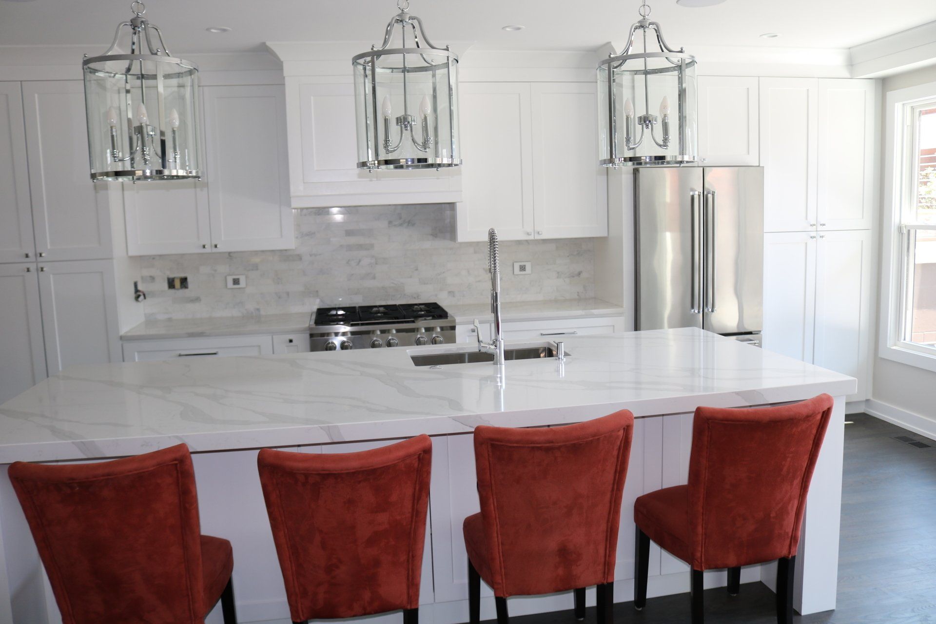 Bright white kitchen with island and orange chairs, stainless steel appliances, and hanging lanterns.