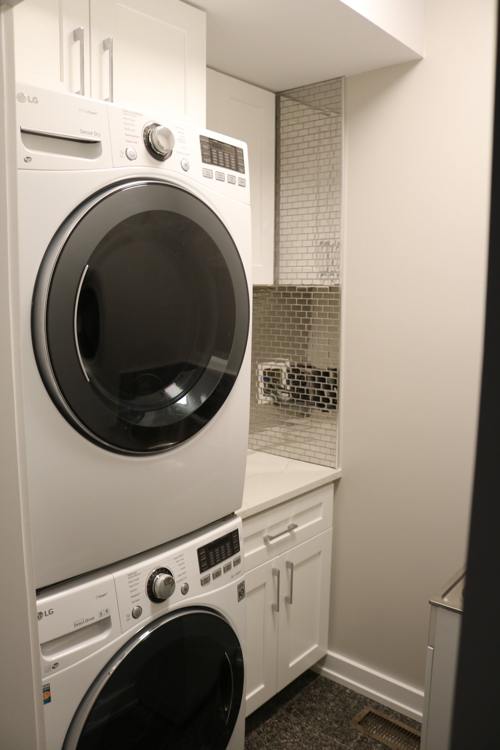 Stacked white LG washer and dryer in a laundry room with white cabinets and a mosaic tile backsplash.