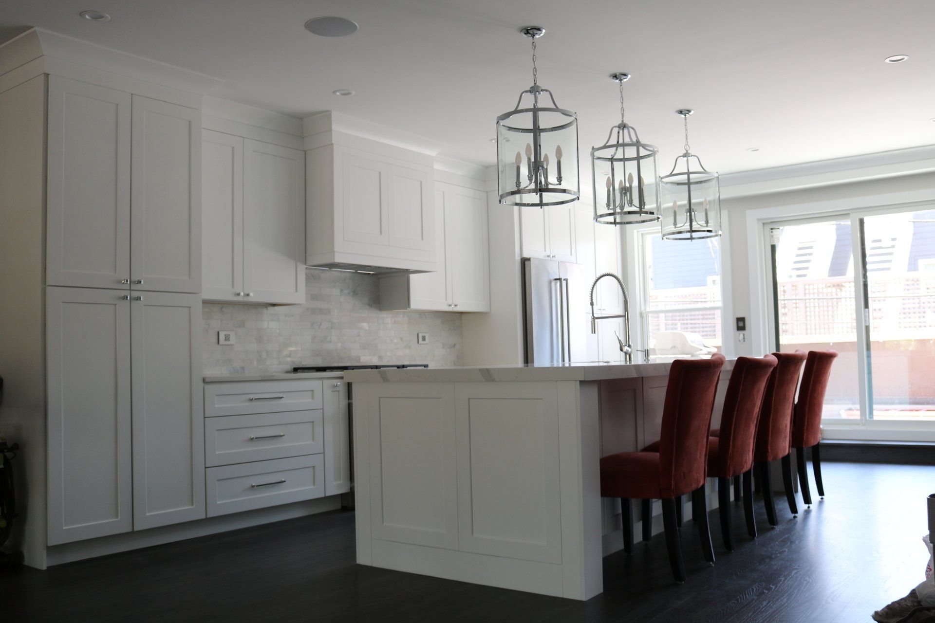 White kitchen with island, three hanging lanterns, and orange chairs. Dark wood floors and sliding glass door.
