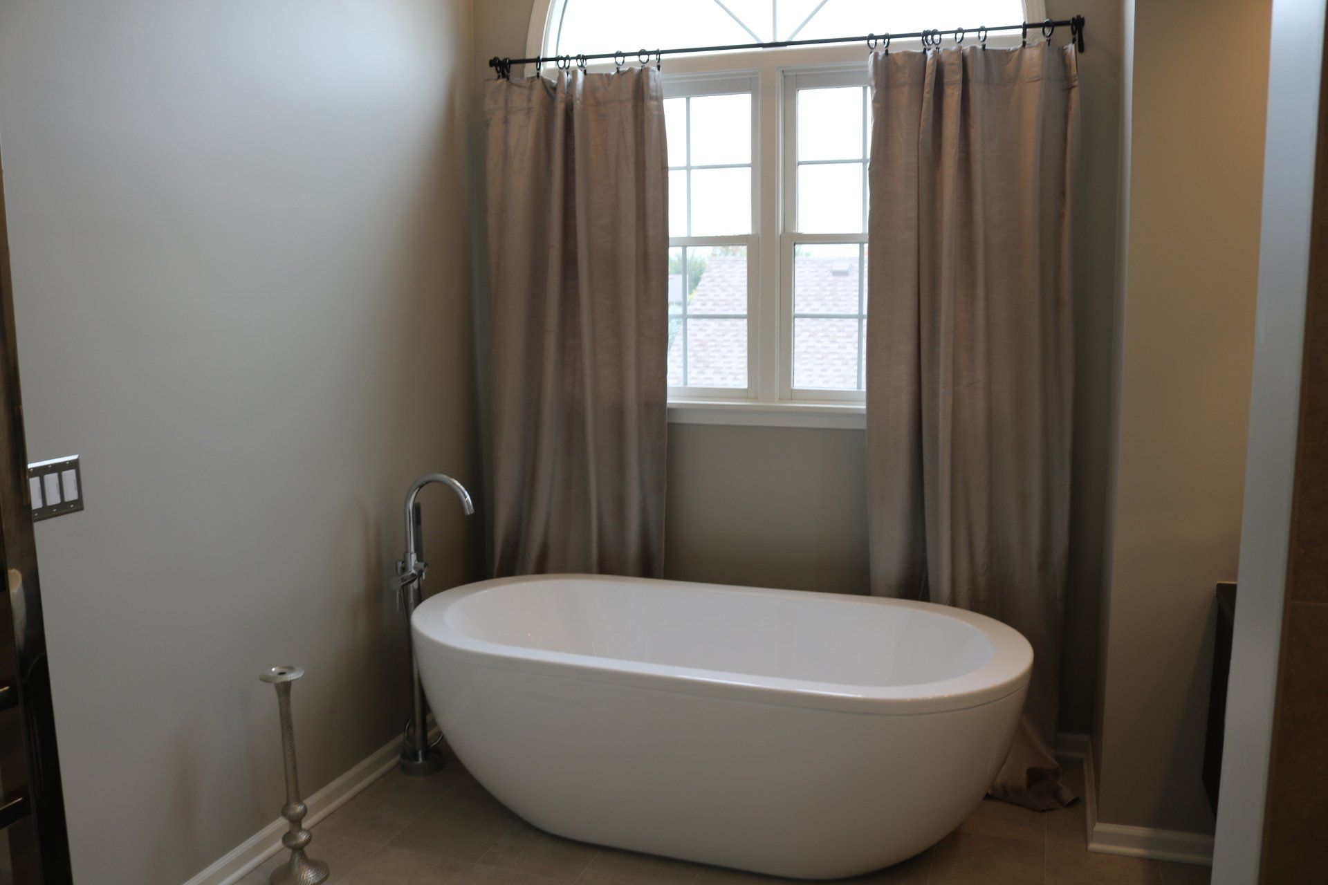 White bathtub in a beige-walled bathroom, beneath a window with beige curtains.