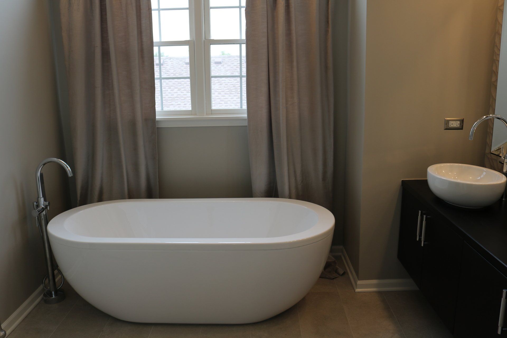Modern bathroom with white soaking tub, grey curtains, and dark vanity with a vessel sink.