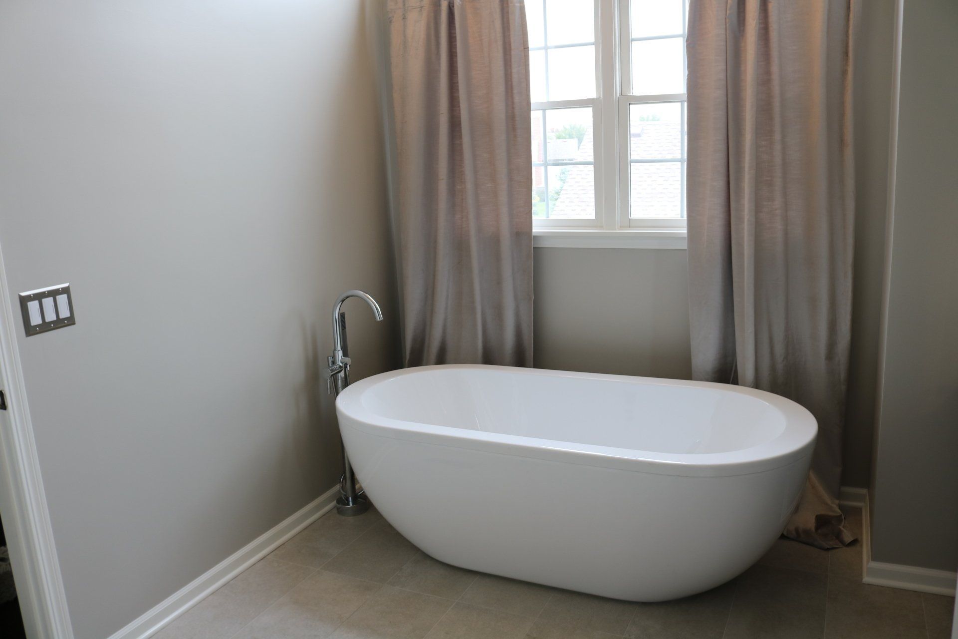 A modern bathroom with a white oval bathtub, light gray walls, and window with curtains.