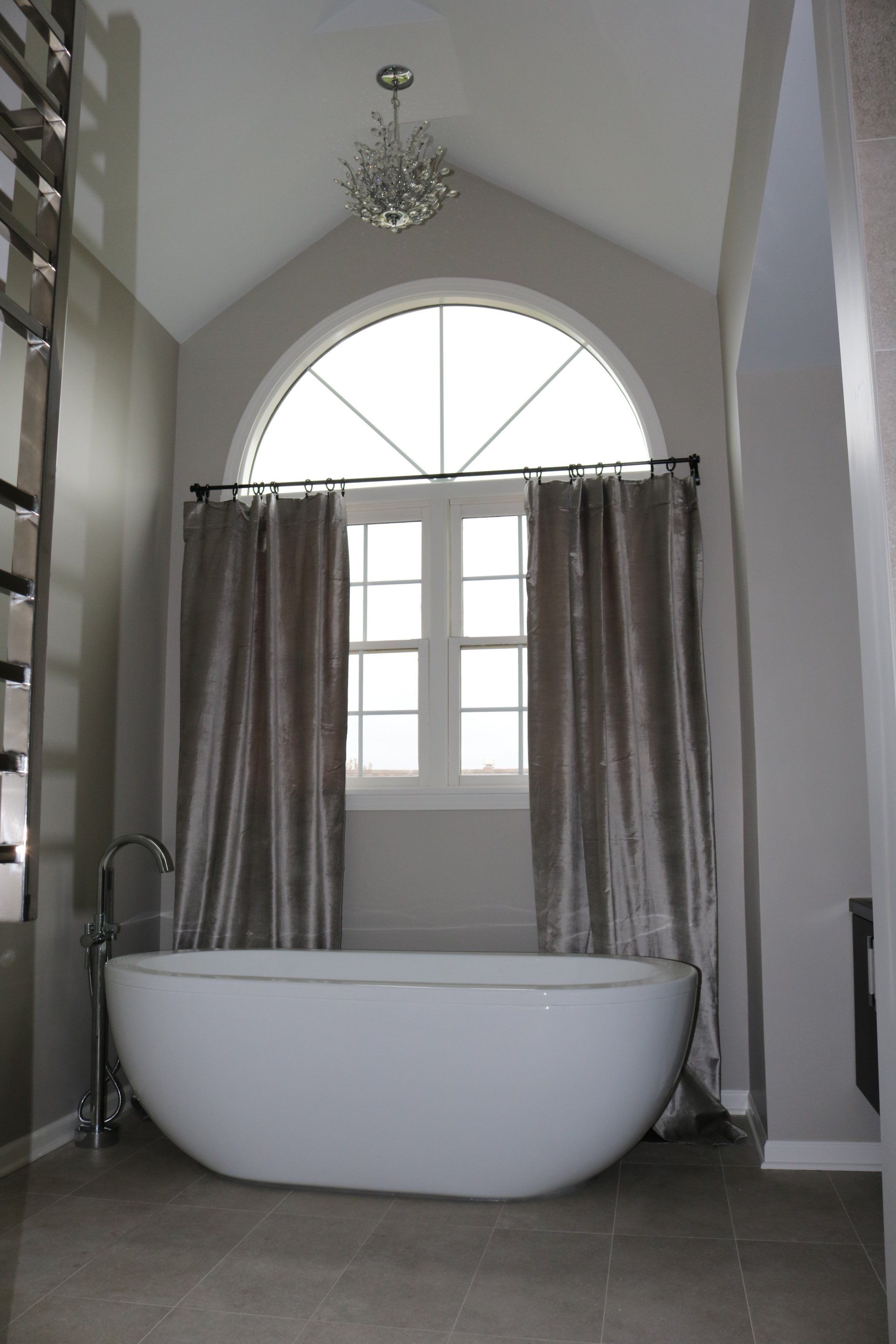 Oval bathtub in a neutral-toned bathroom with a large arched window, curtains, and a chandelier.