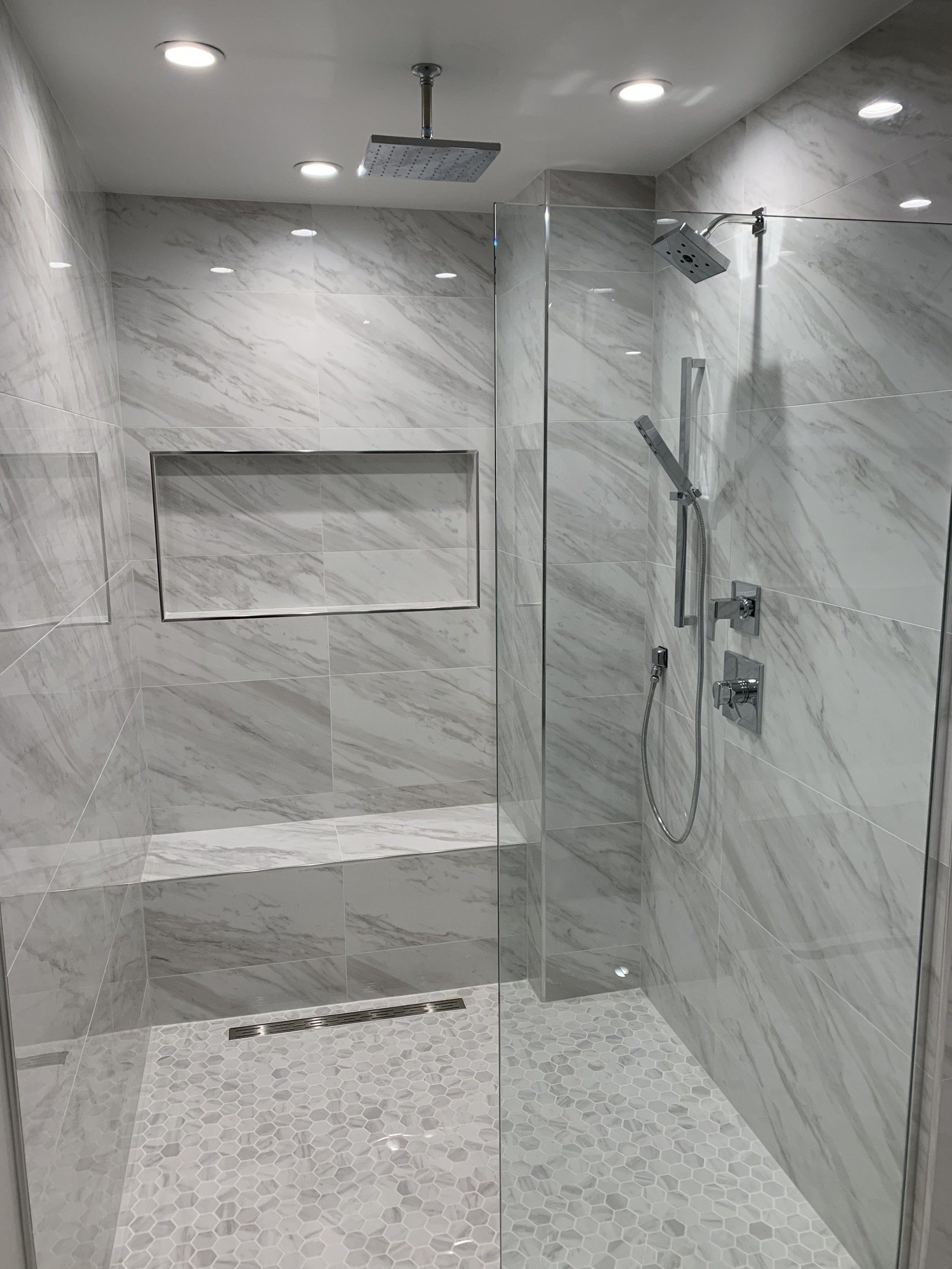 Modern white tiled shower with glass door and built-in bench, overhead and handheld shower heads.