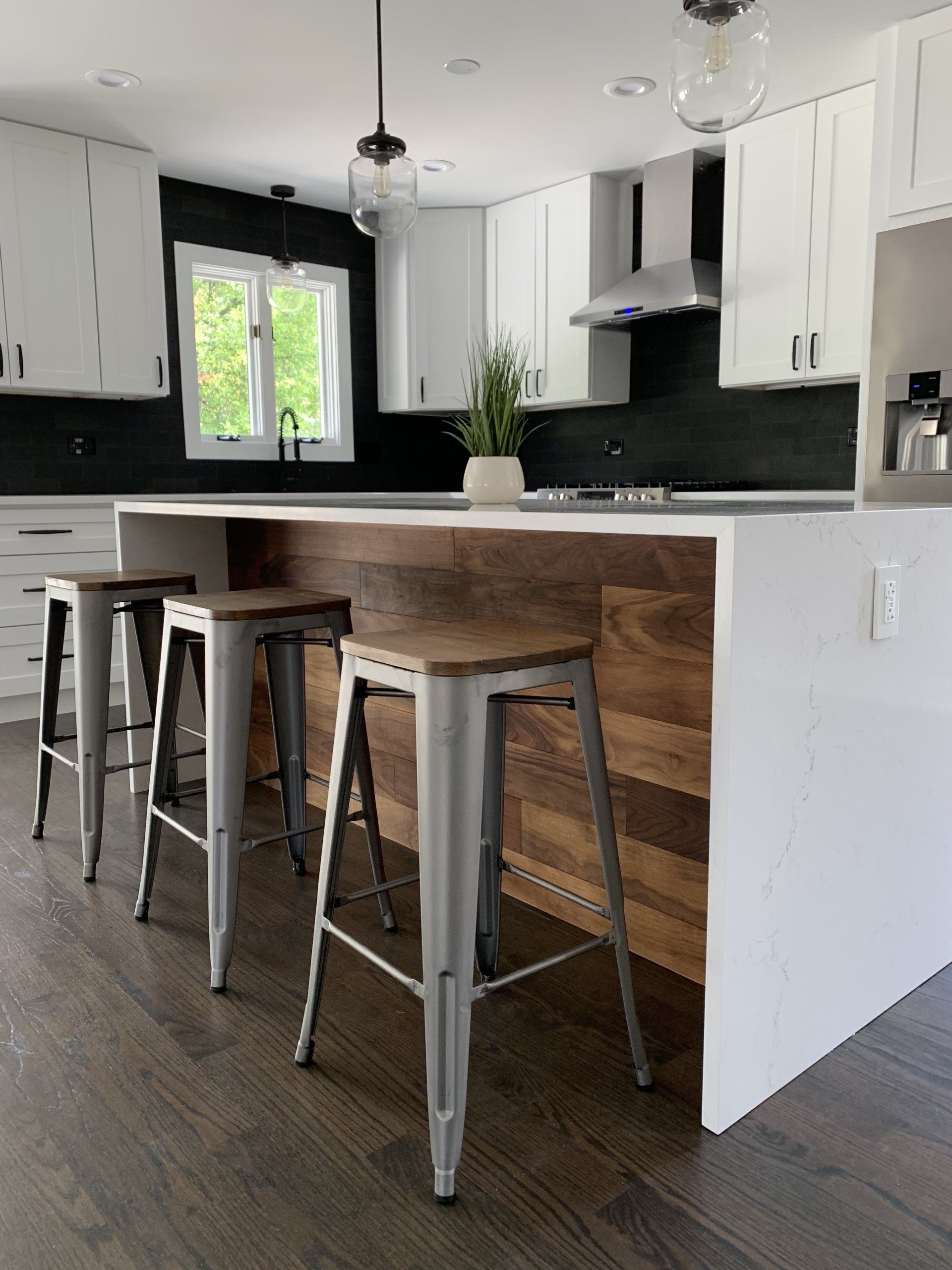 Modern kitchen with white cabinets, wooden island, bar stools, dark backsplash, and wood floor.