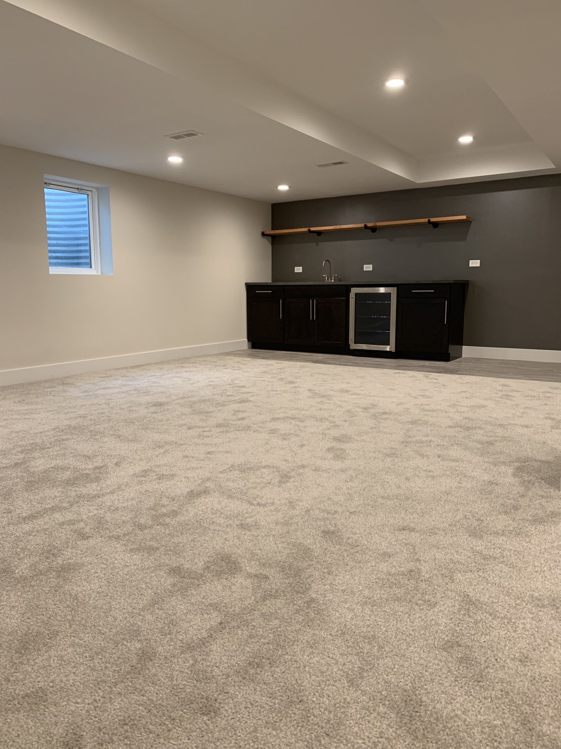 Basement with dark bar, mini-fridge, and shelves against a gray accent wall. Light carpet and beige walls.