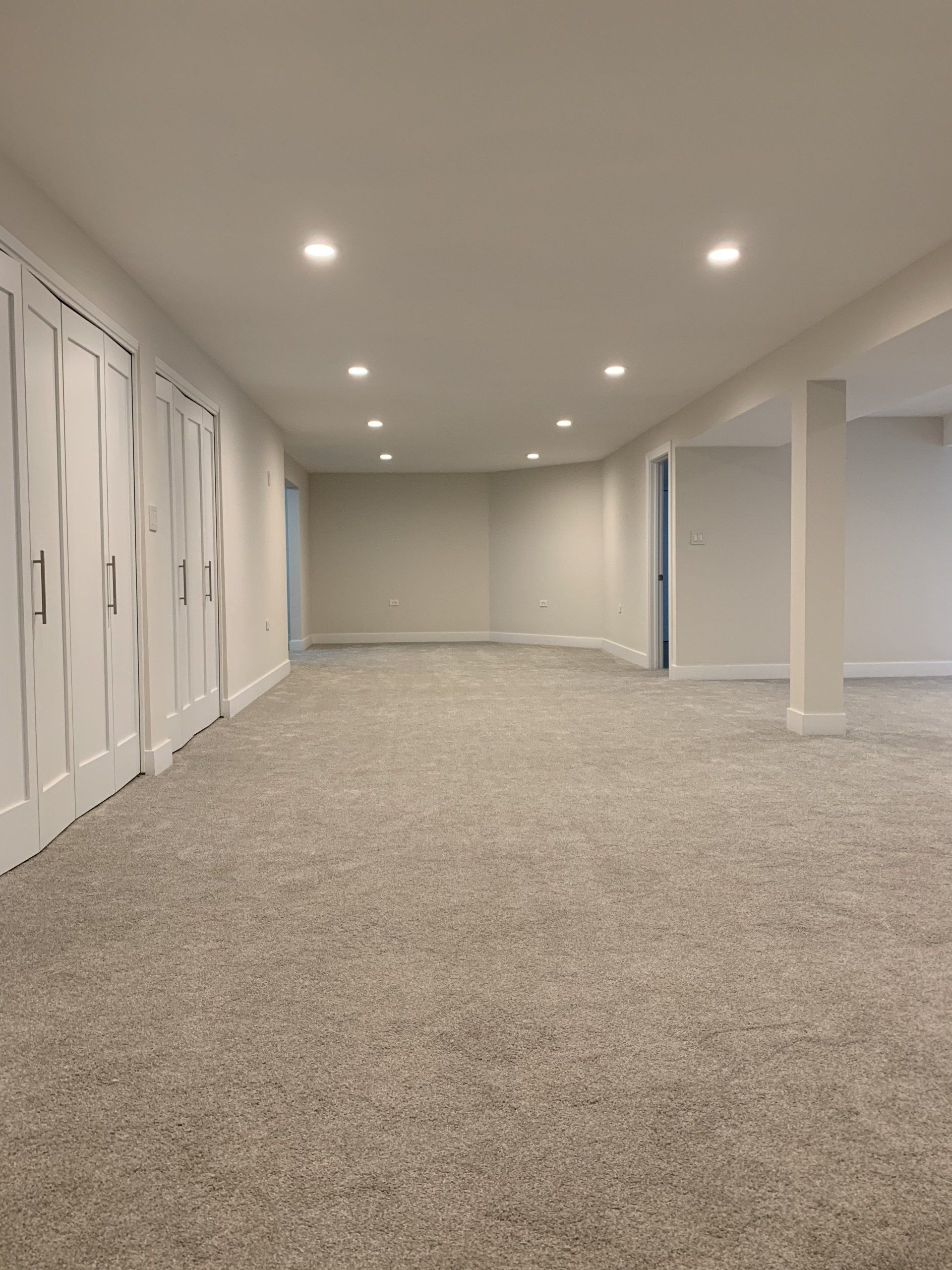 Empty, white-walled room with recessed lighting and light gray speckled carpet. There are white doors on the left and support columns on the right.