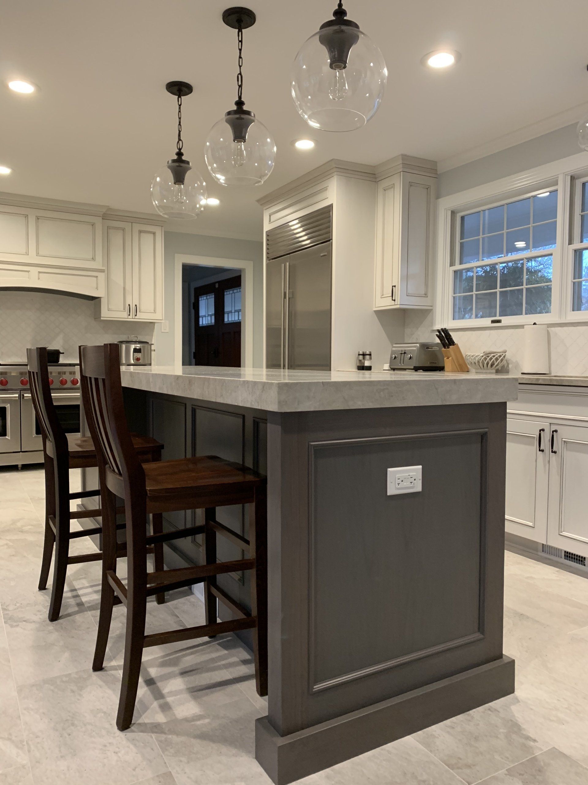 Kitchen with gray island, white cabinets, wooden bar stools, and pendant lights.