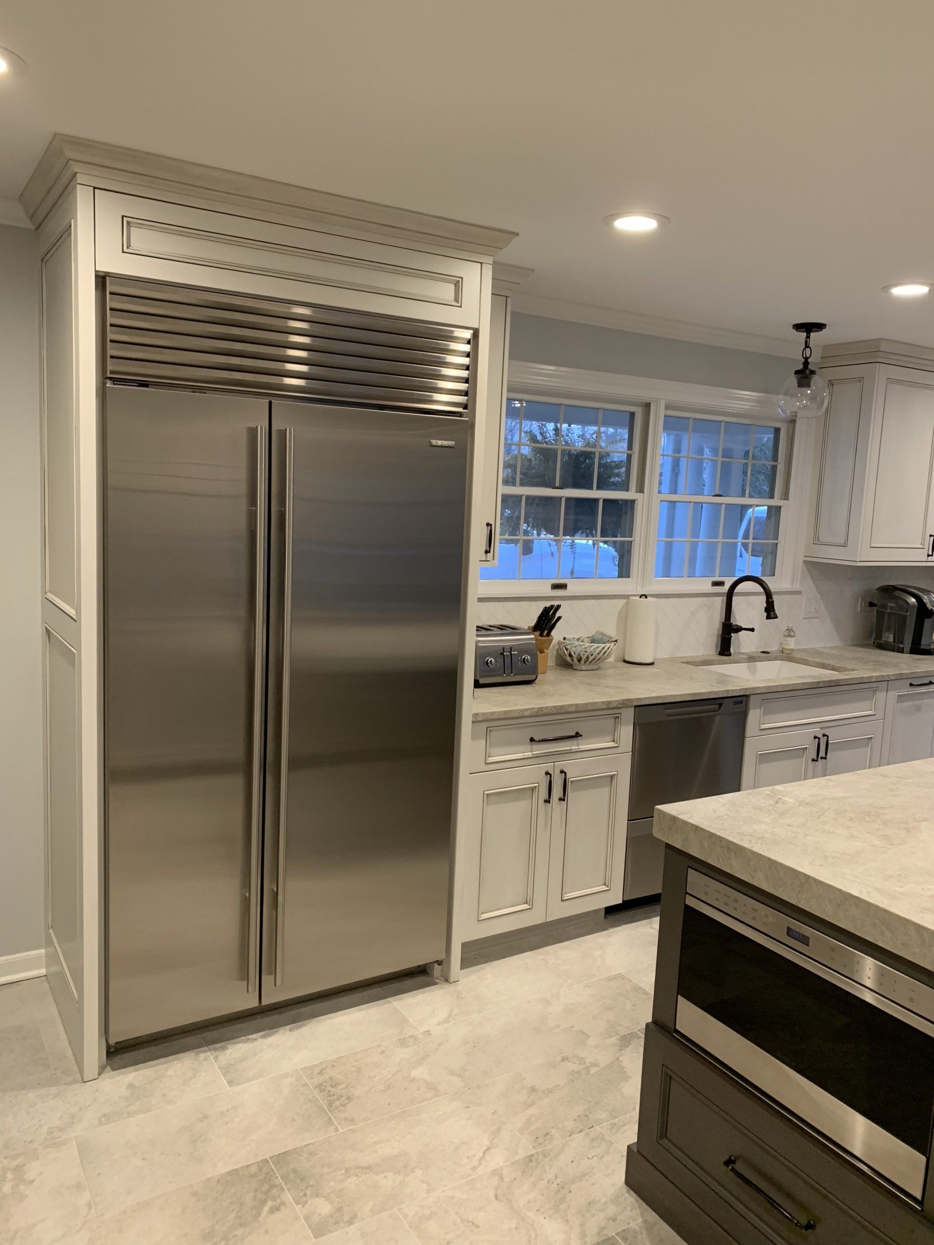 Stainless steel refrigerator in white kitchen; light gray floors, window, sink, island with oven.