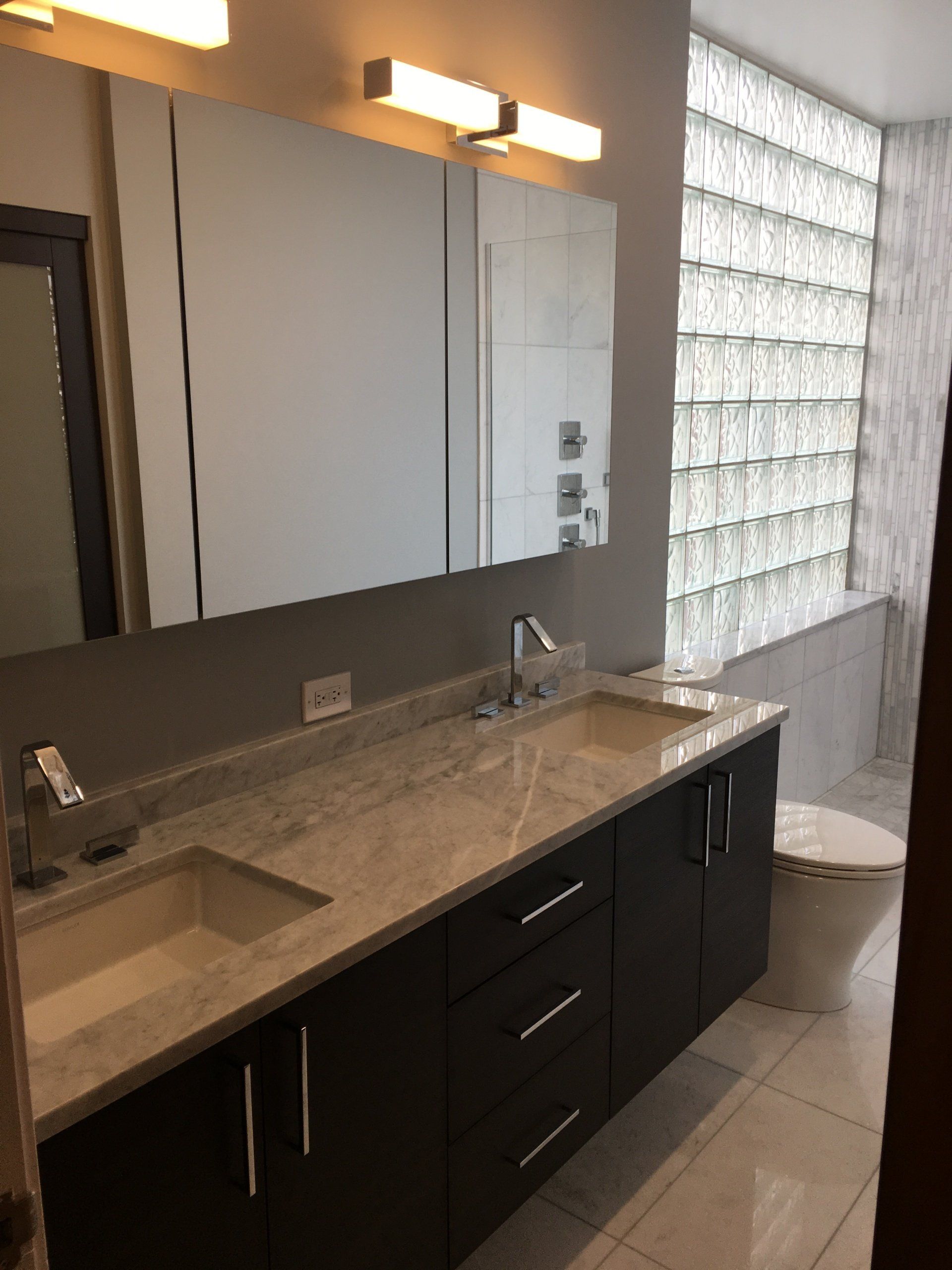 Modern bathroom with dark vanity, dual sinks, large mirror, and glass block window.