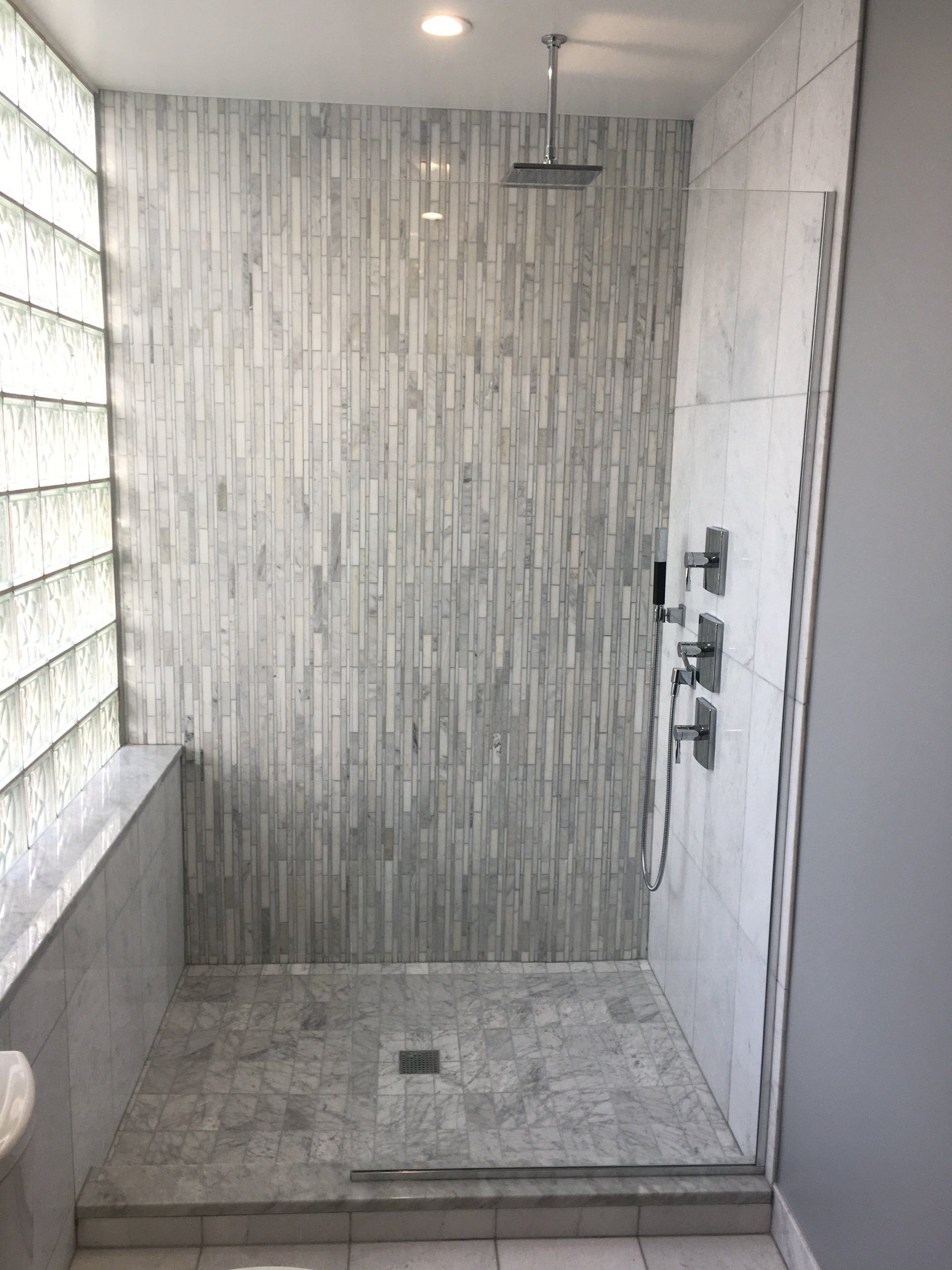 A modern shower with gray tiled walls, glass block window, and chrome fixtures.