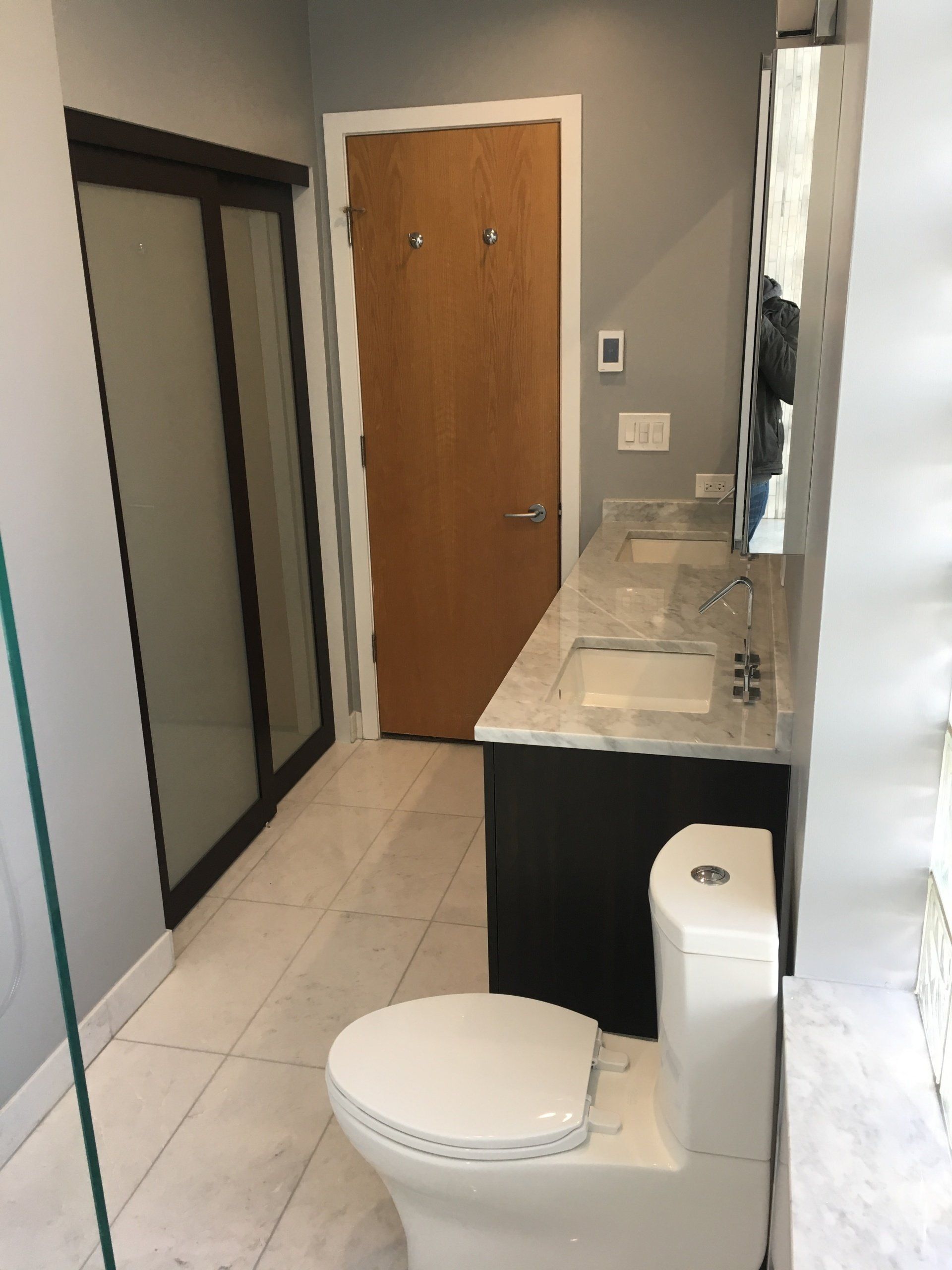 Bathroom with toilet, vanity, and shower. Gray walls, white floor, and brown door.