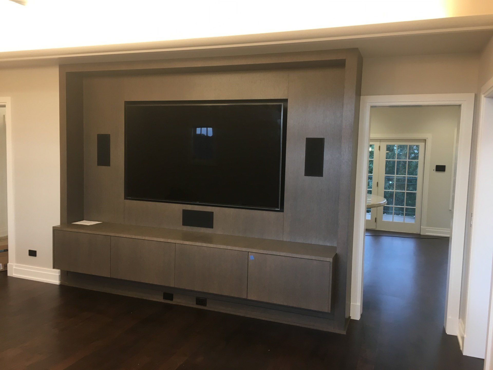 A large TV centered in a custom wooden entertainment center. Speakers and floating cabinets present. Dark wood floors.