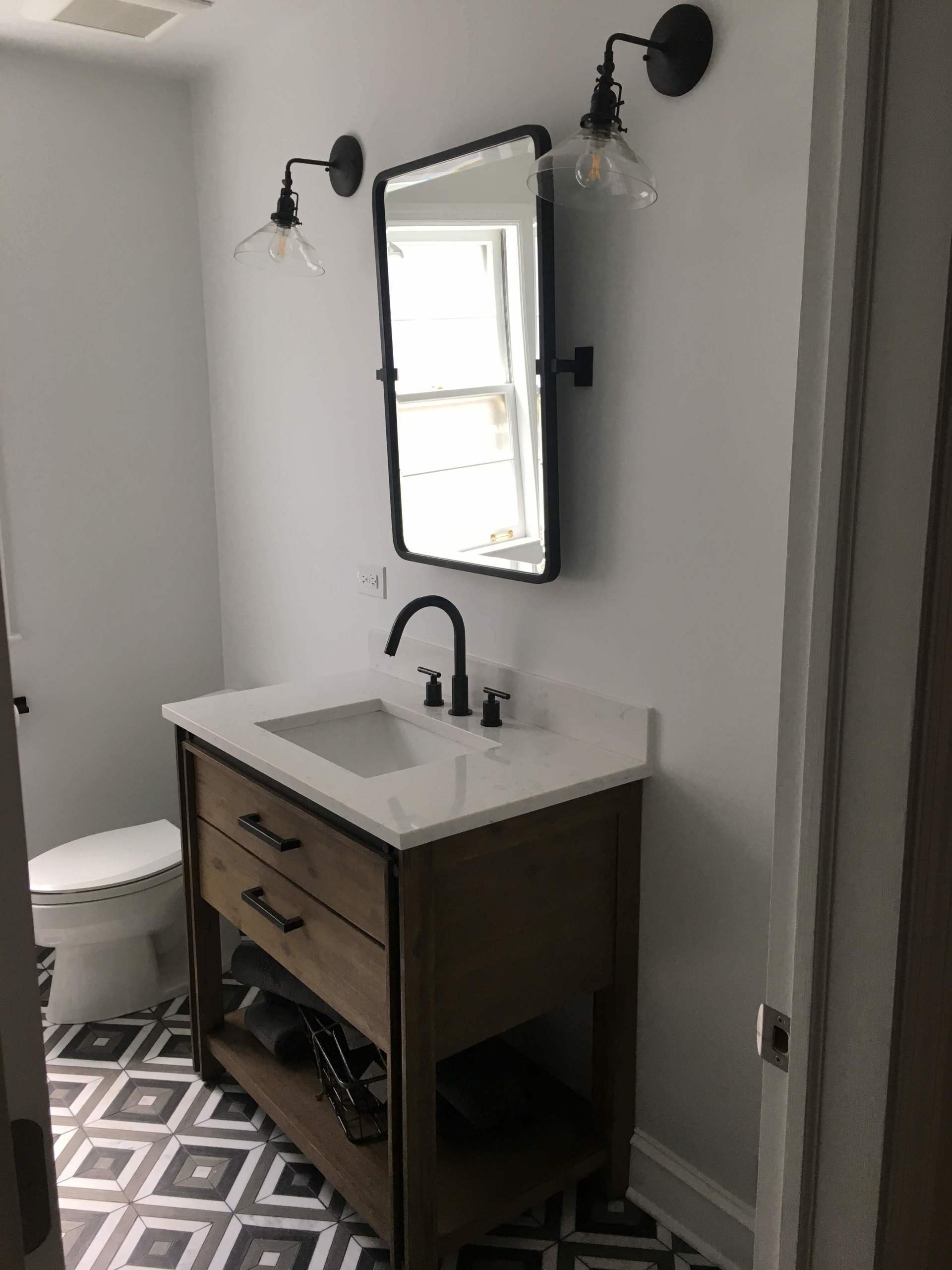 Bathroom with wood vanity, black faucet, mirror, sconces, and patterned tile floor.