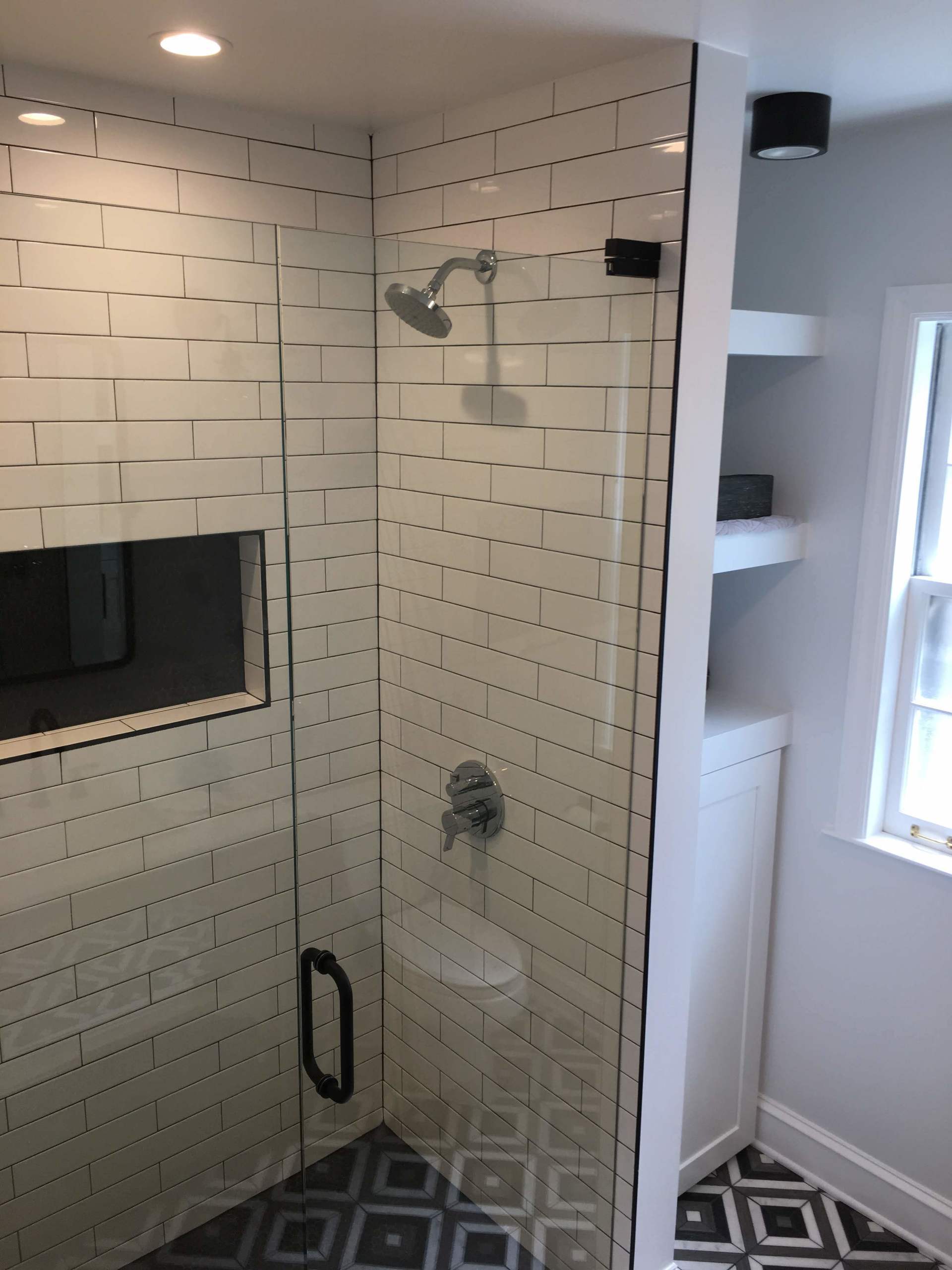 White-tiled shower with glass door, rain shower head, and black accents; built-in shelves and window beside it.