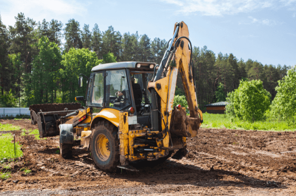 Yellow backhoe on a dirt field, with forest in background and a clear sky.