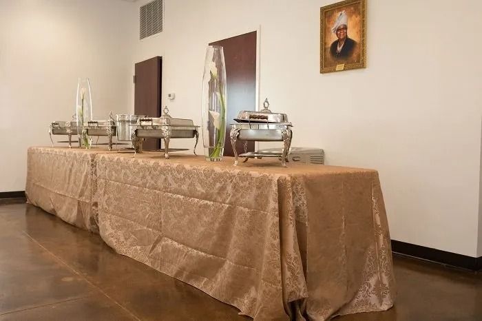 A buffet table with chafing dishes and decorative glass vases draped in a gold tablecloth in a room with a framed portrait.