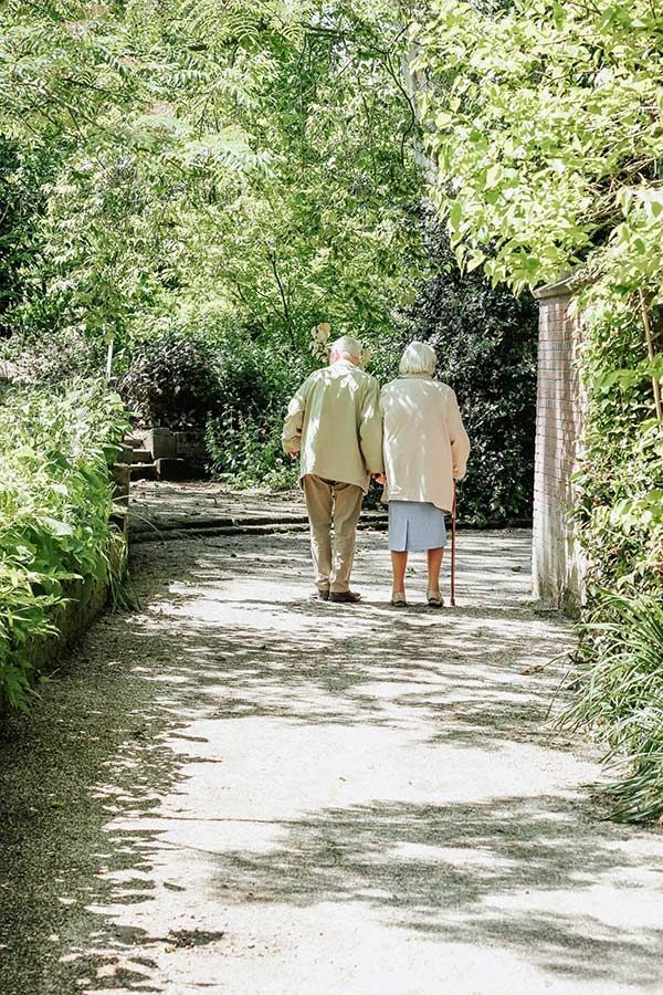 Two people walk away from the viewer along a sun-dappled path in a lush, green wooded area.