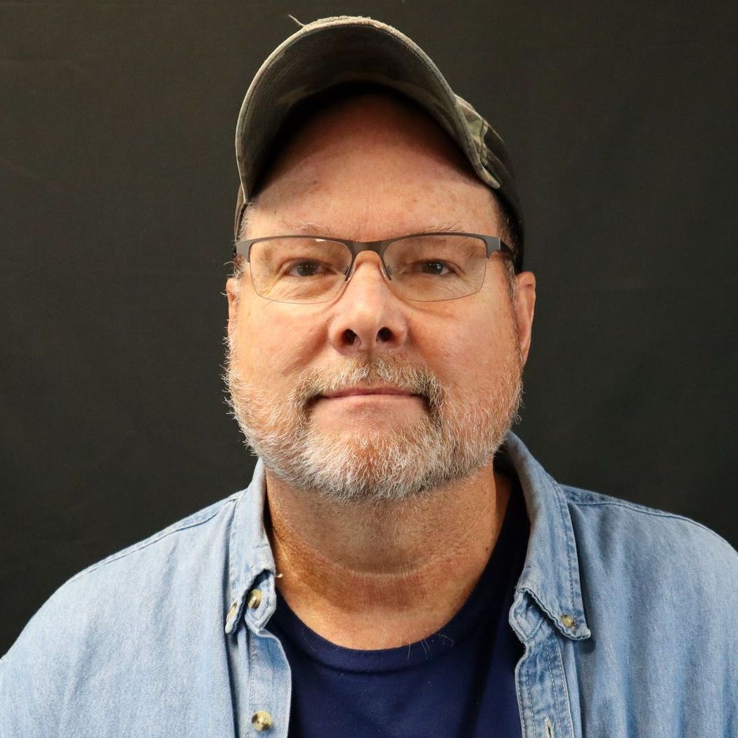 A man with a beard and glasses is wearing a hat and a blue shirt.