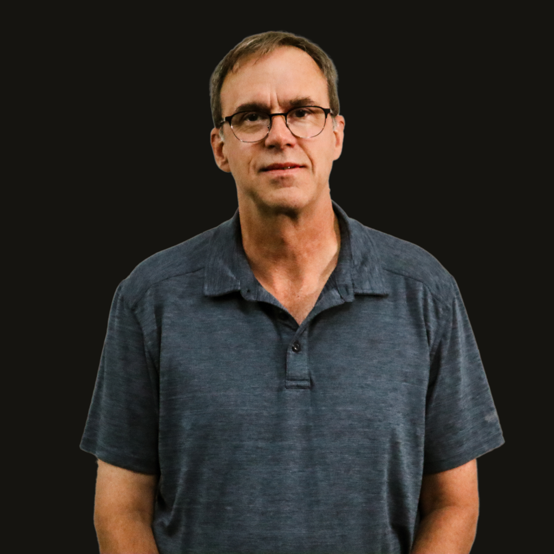 A man wearing glasses and a blue shirt is standing in front of a black background.