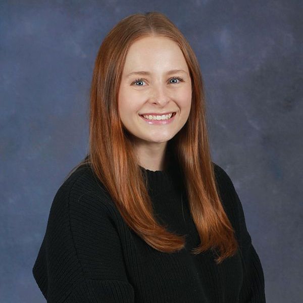 A woman with long red hair is wearing a grey sweater and smiling.