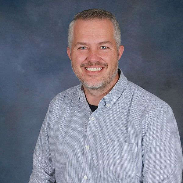 A man with a beard is smiling for the camera while wearing a black sweater and a blue shirt.