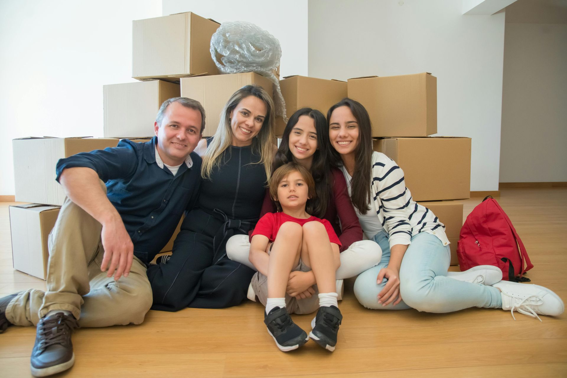 Family of five sits on floor surrounded by moving boxes; smiling faces, new home.