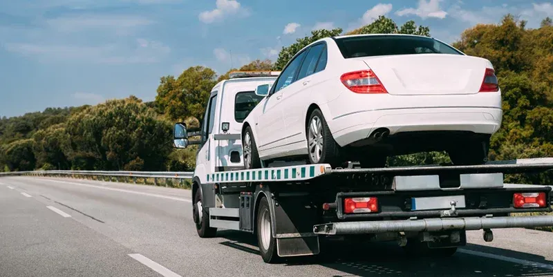 White car being towed on a flatbed tow truck on a highway. Sunny day, trees in the background.