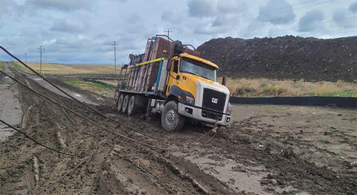 Yellow dump truck stuck in muddy road, tilting to the side. Overcast sky and pile of earth in background.