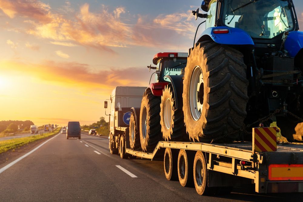 A trailer carrying two tractors driving on a highway at sunset.