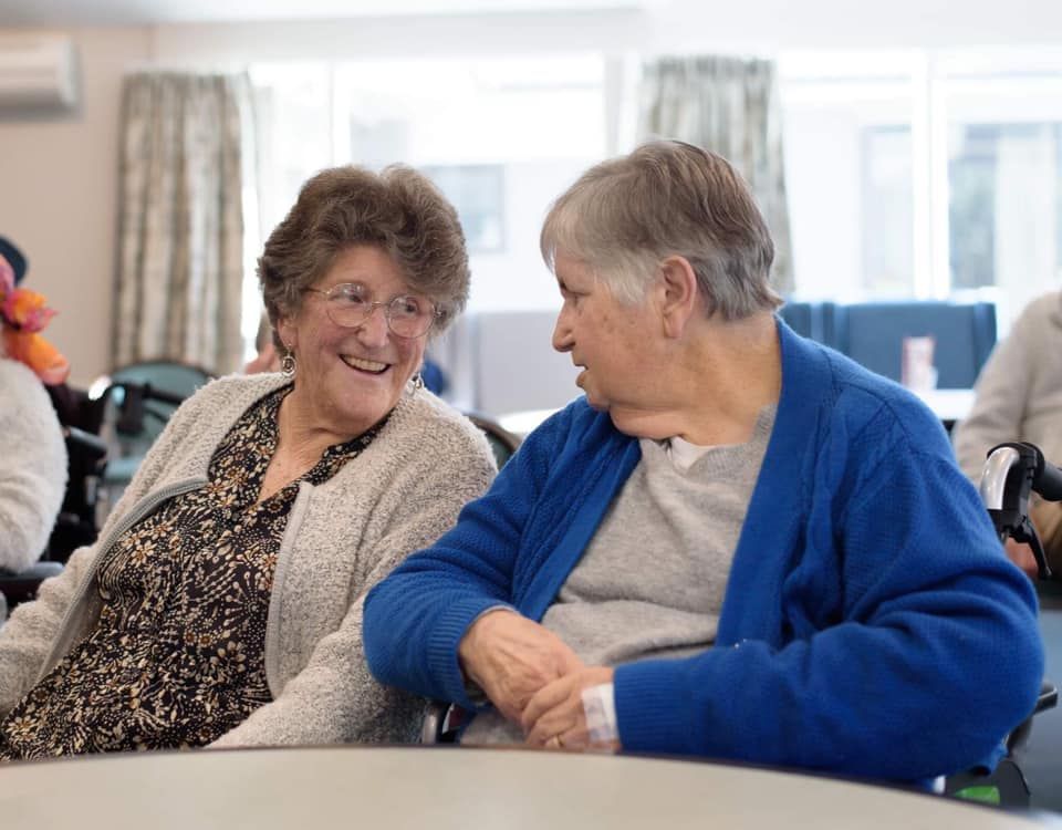 Two elderly women are sitting at a table talking to each other.