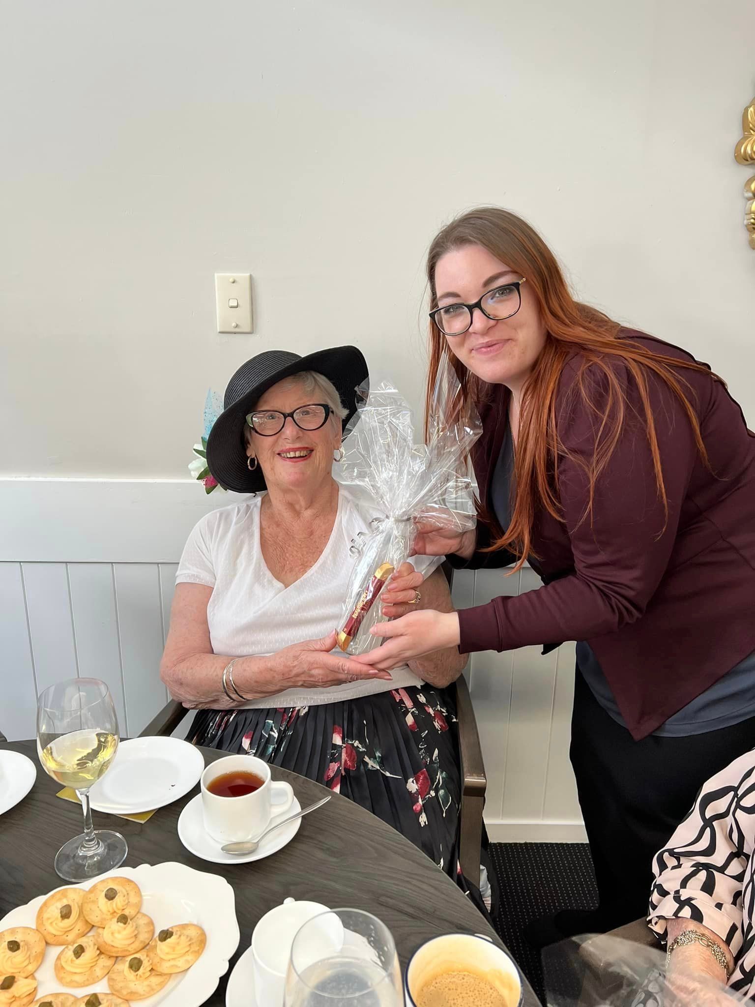 A woman is giving a gift to an elderly woman sitting at a table.