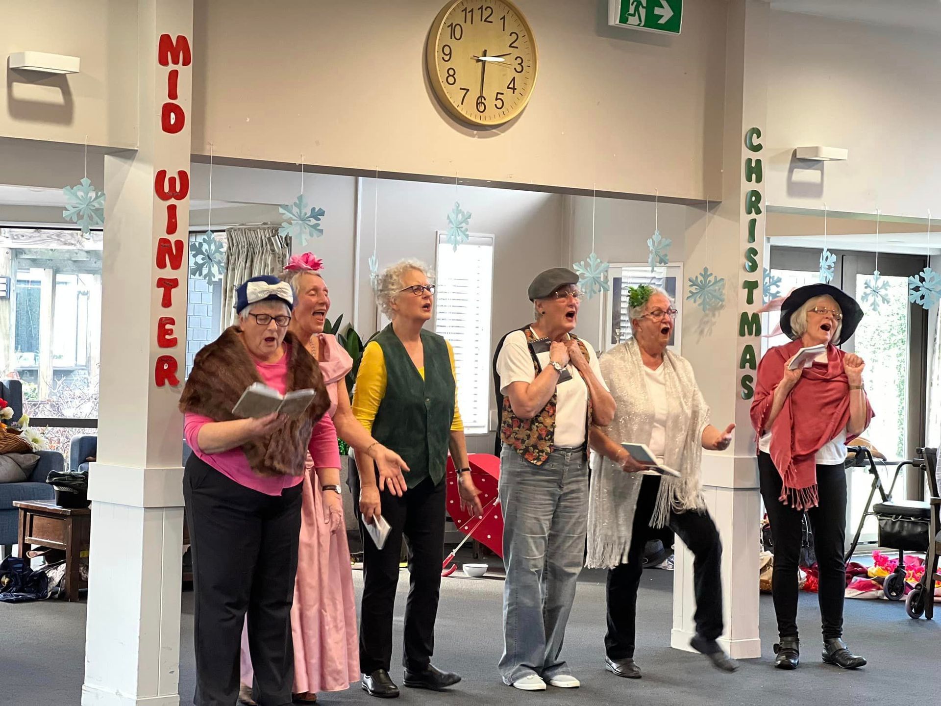 A group of women are singing in front of a clock in a room.