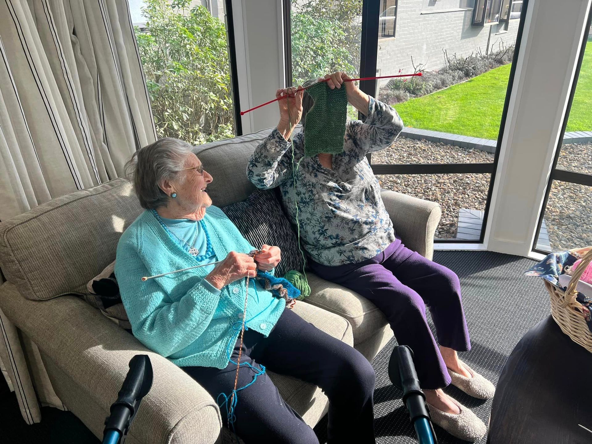 Two elderly women are sitting on a couch knitting.