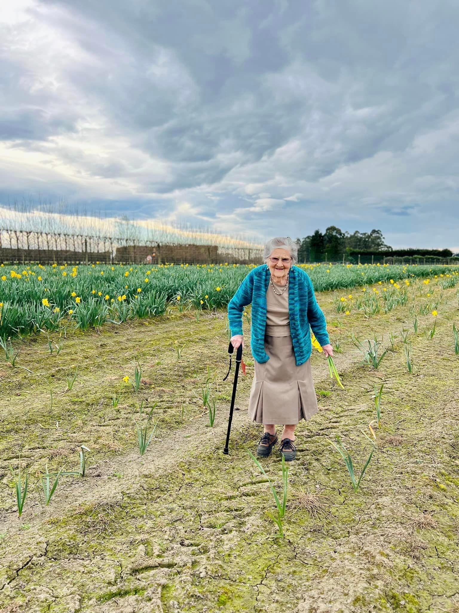 An elderly woman is walking through a field of flowers.