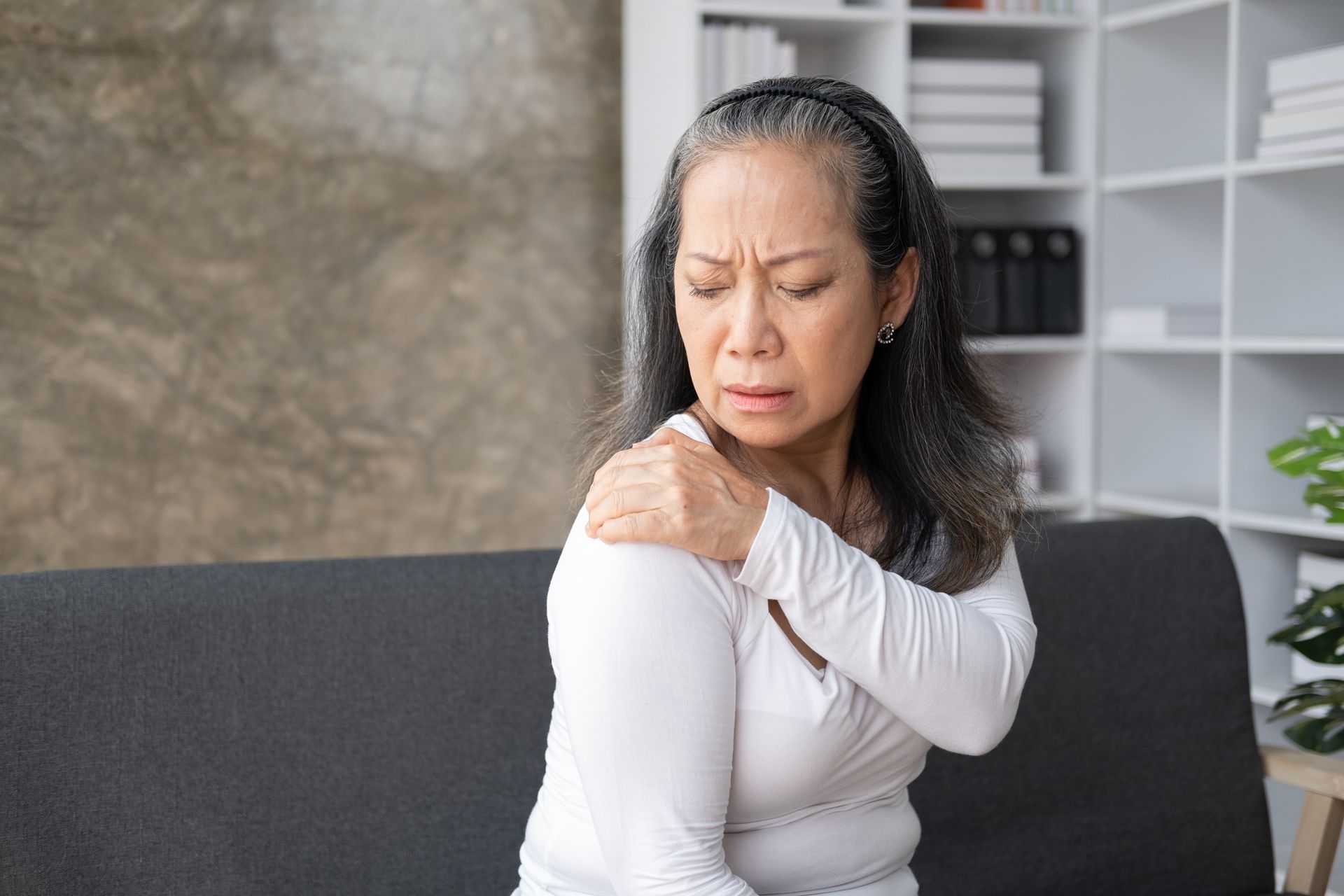 Uma mulher mais velha está sentada em um sofá segurando o ombro com dor.