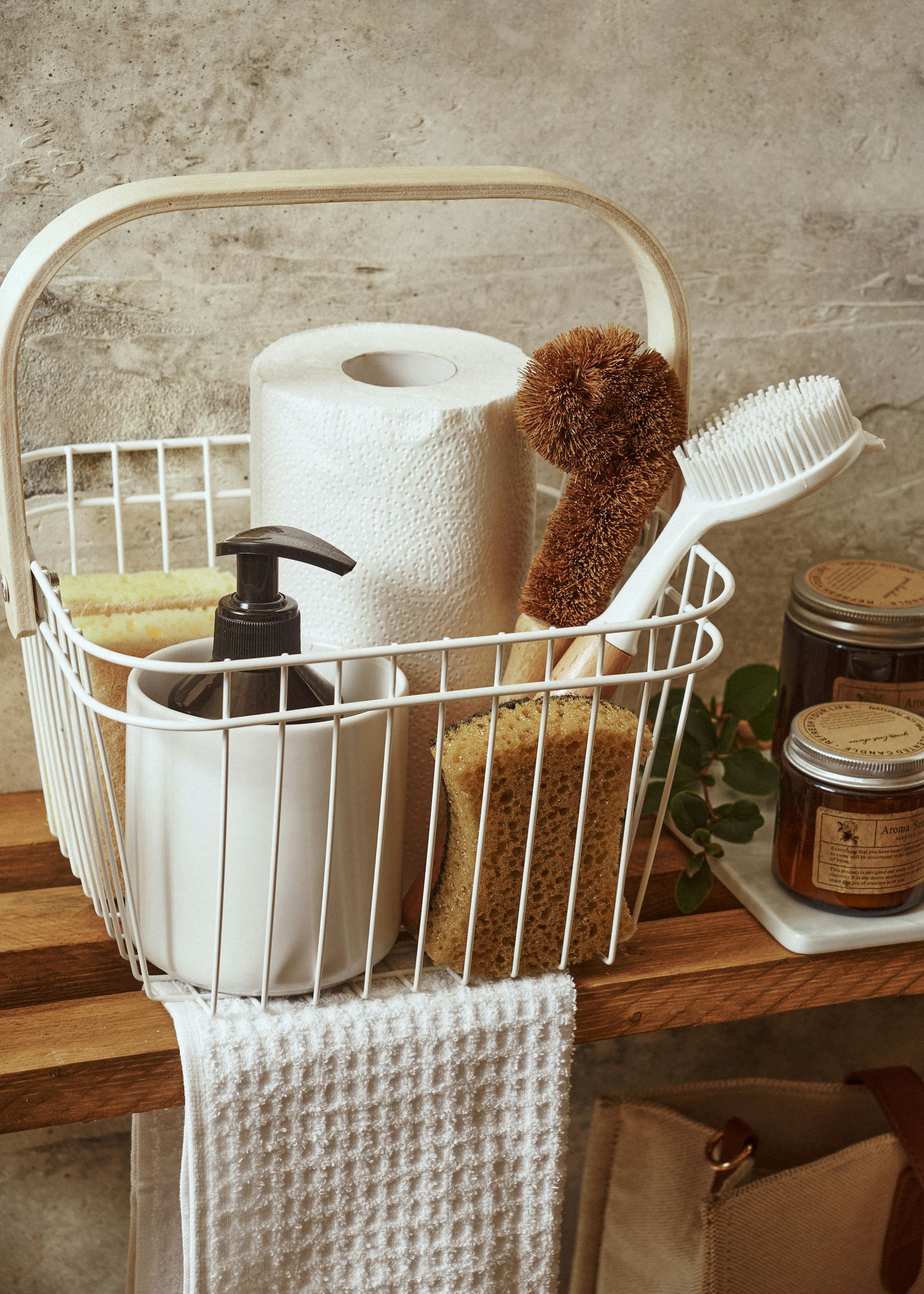 White wire basket holding toiletries: toilet paper, soap dispenser, sponge, brush, and a wooden handle.