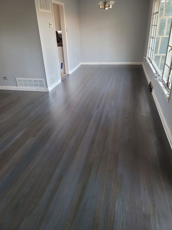 Empty modern room with gray wood flooring, white walls, and large windows along the right side.