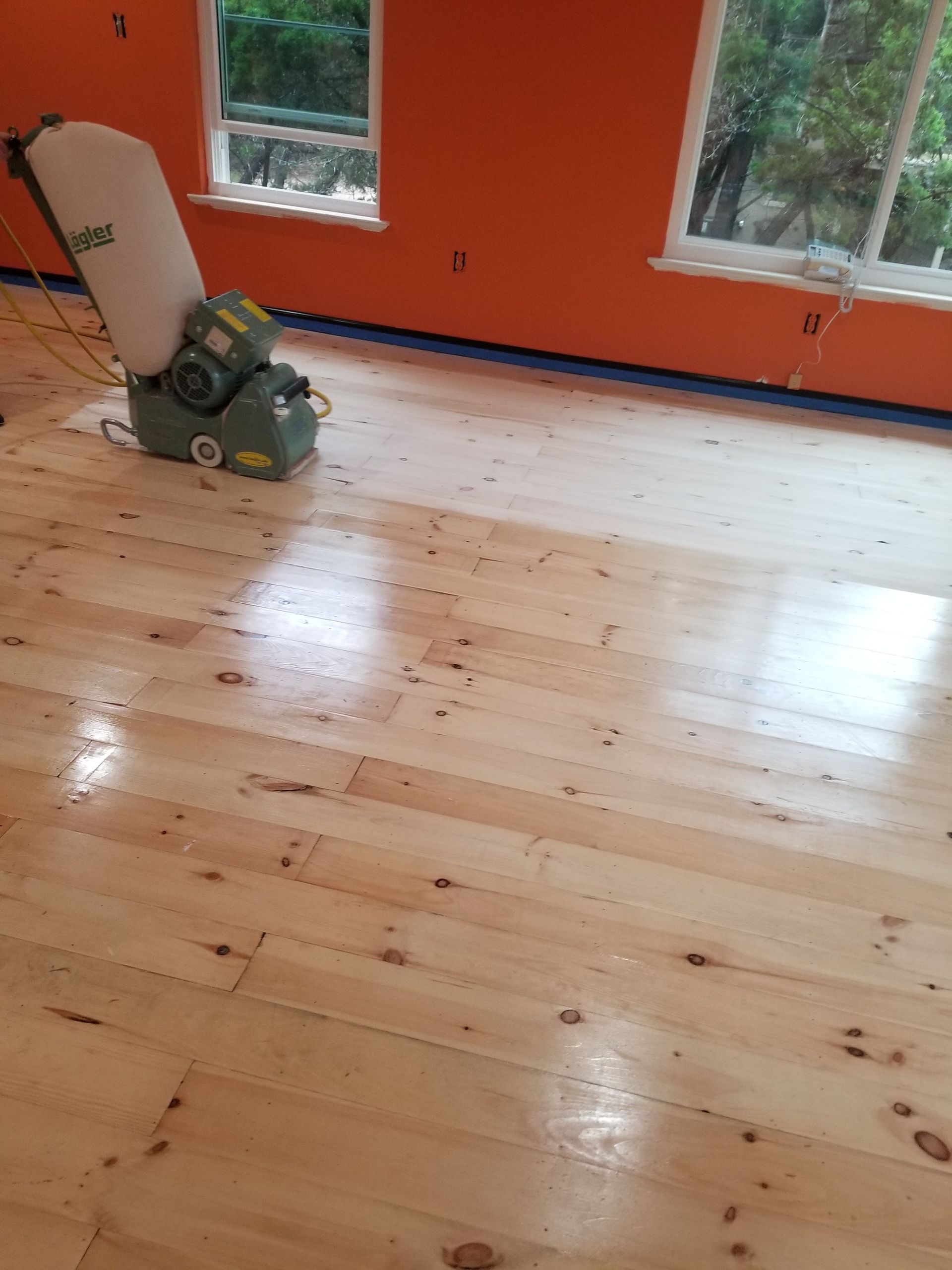 Empty room with freshly sanded hardwood floor and a floor sander near orange walls and windows