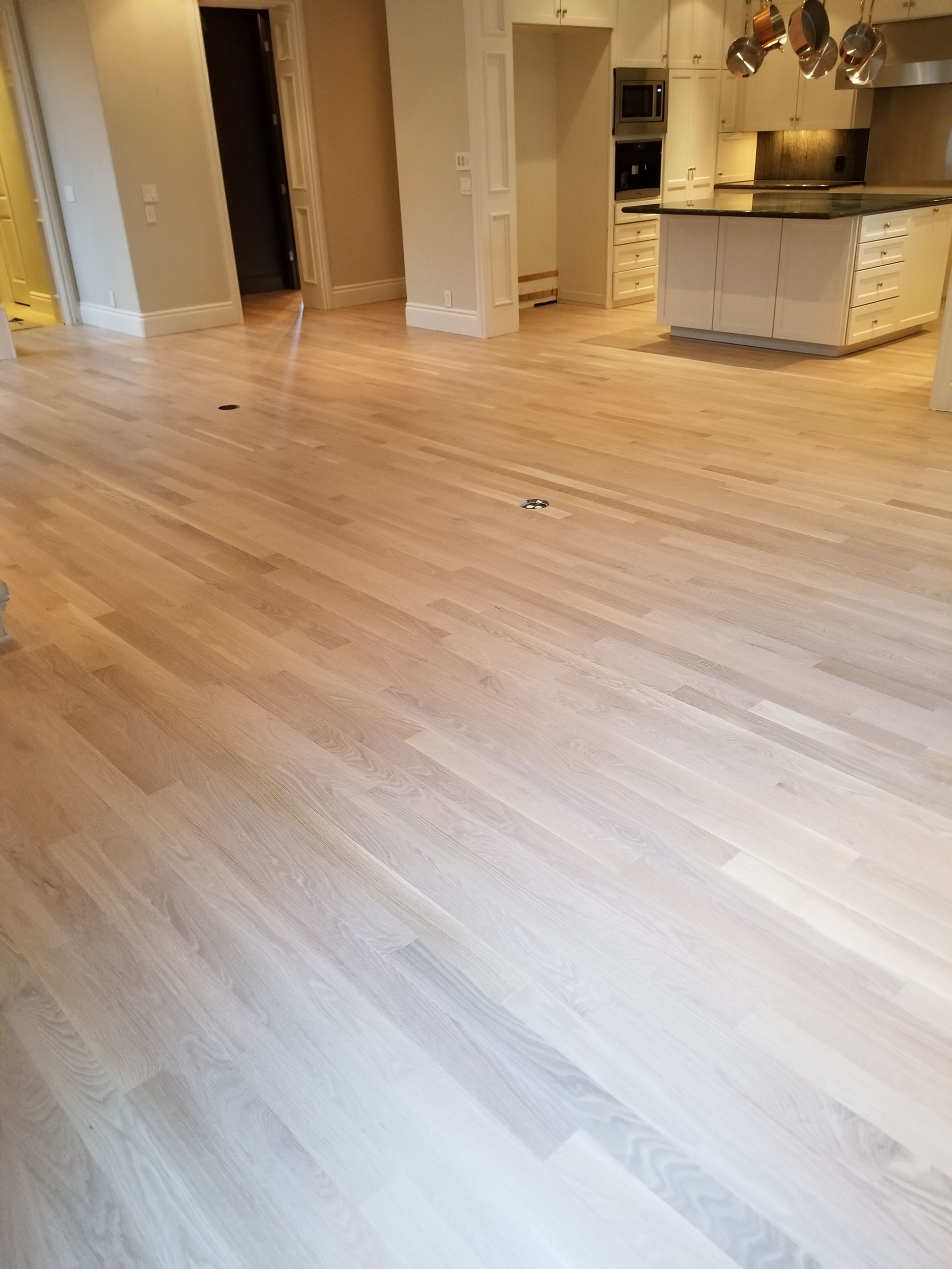 Empty light wood-floored room with white columns, open doorways, and a kitchen island in the background