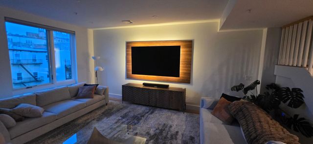 A modern living room with a large TV mounted on a wood-paneled wall, two sofas, and a large window.
